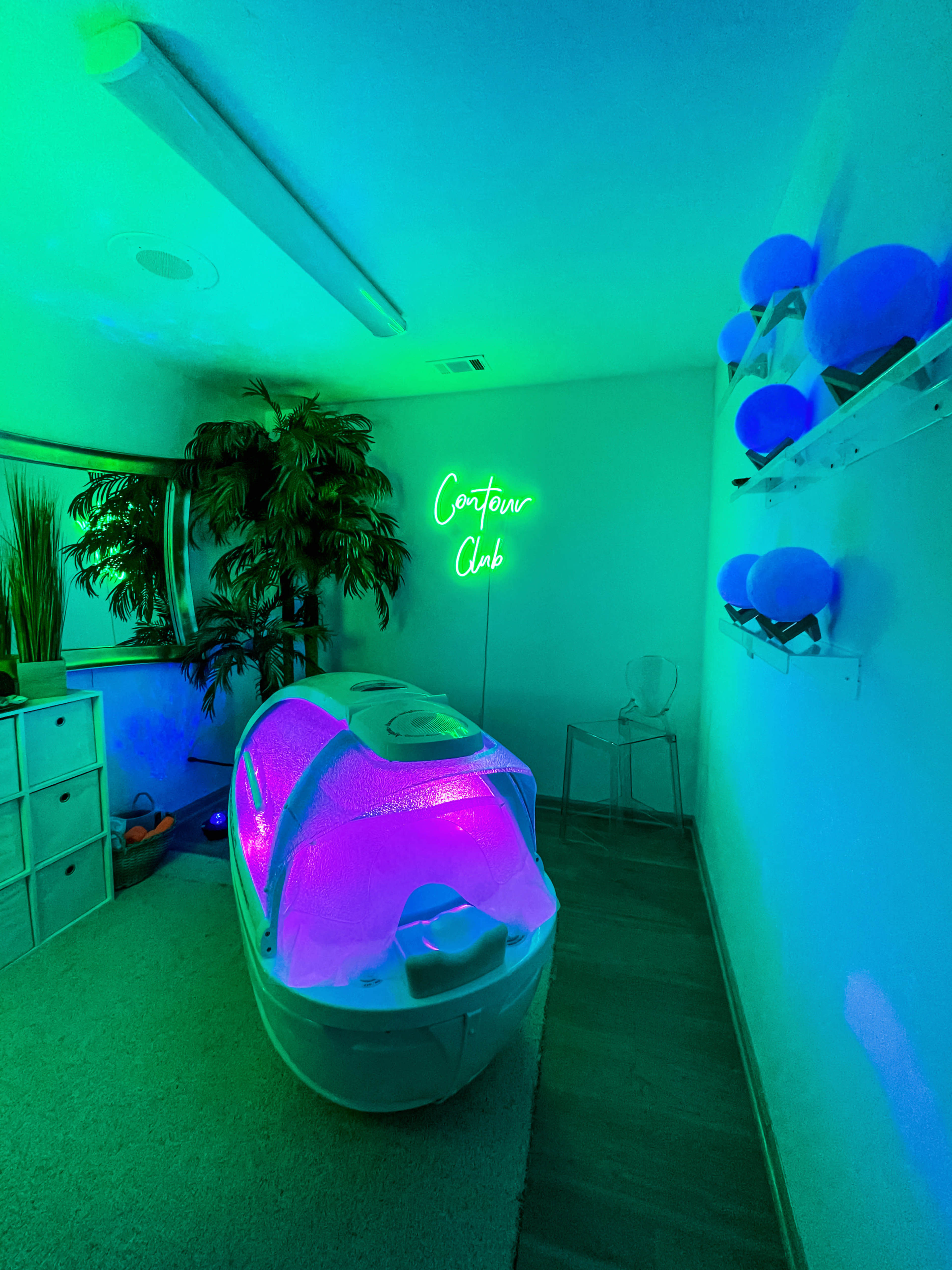 A spa room with a light therapy machine covered in a glowing purple cover, surrounded by green lighting and decorative plants.