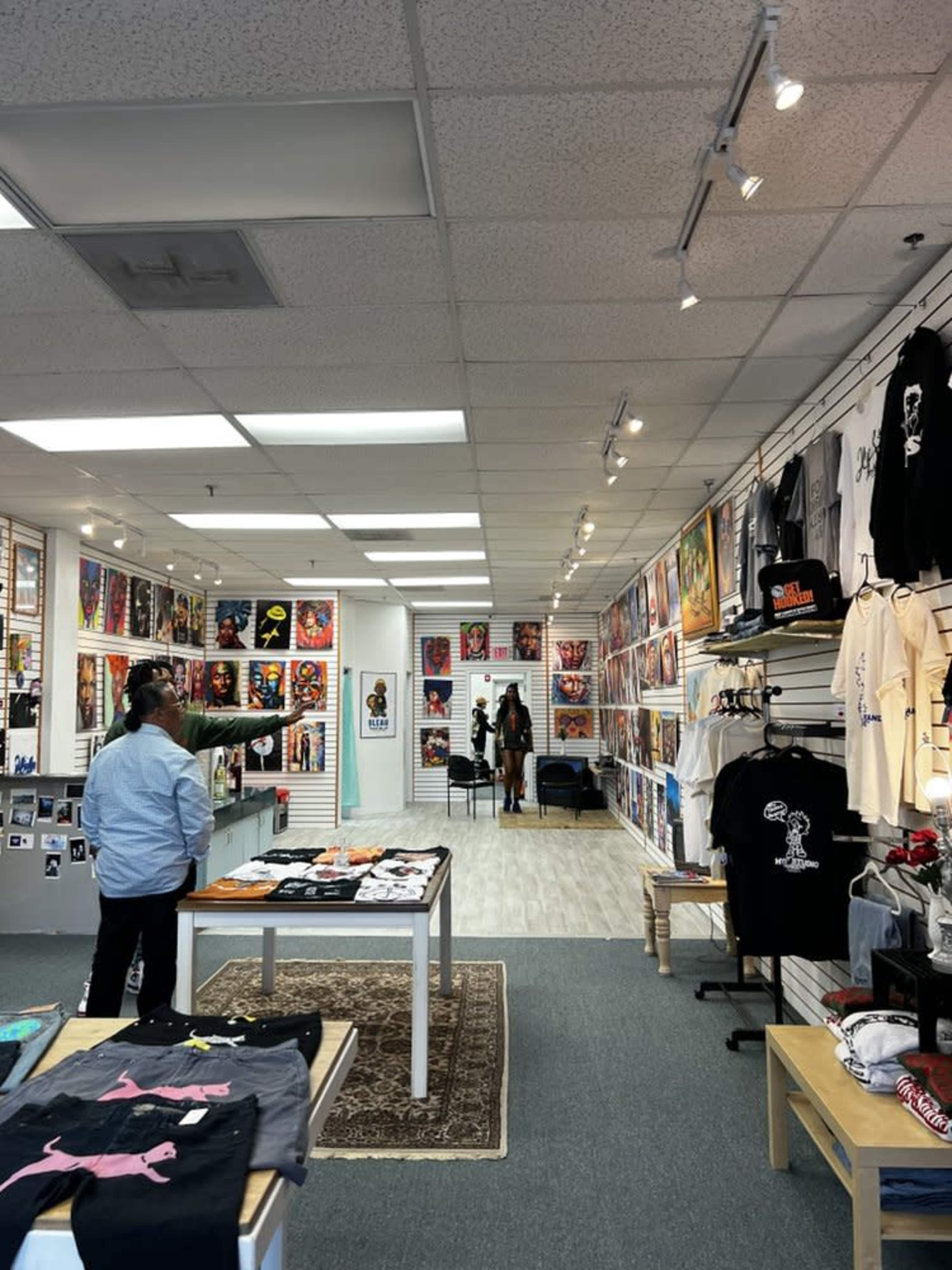 The image shows an interior view of a retail store displaying various artworks and apparel on the walls and tables.