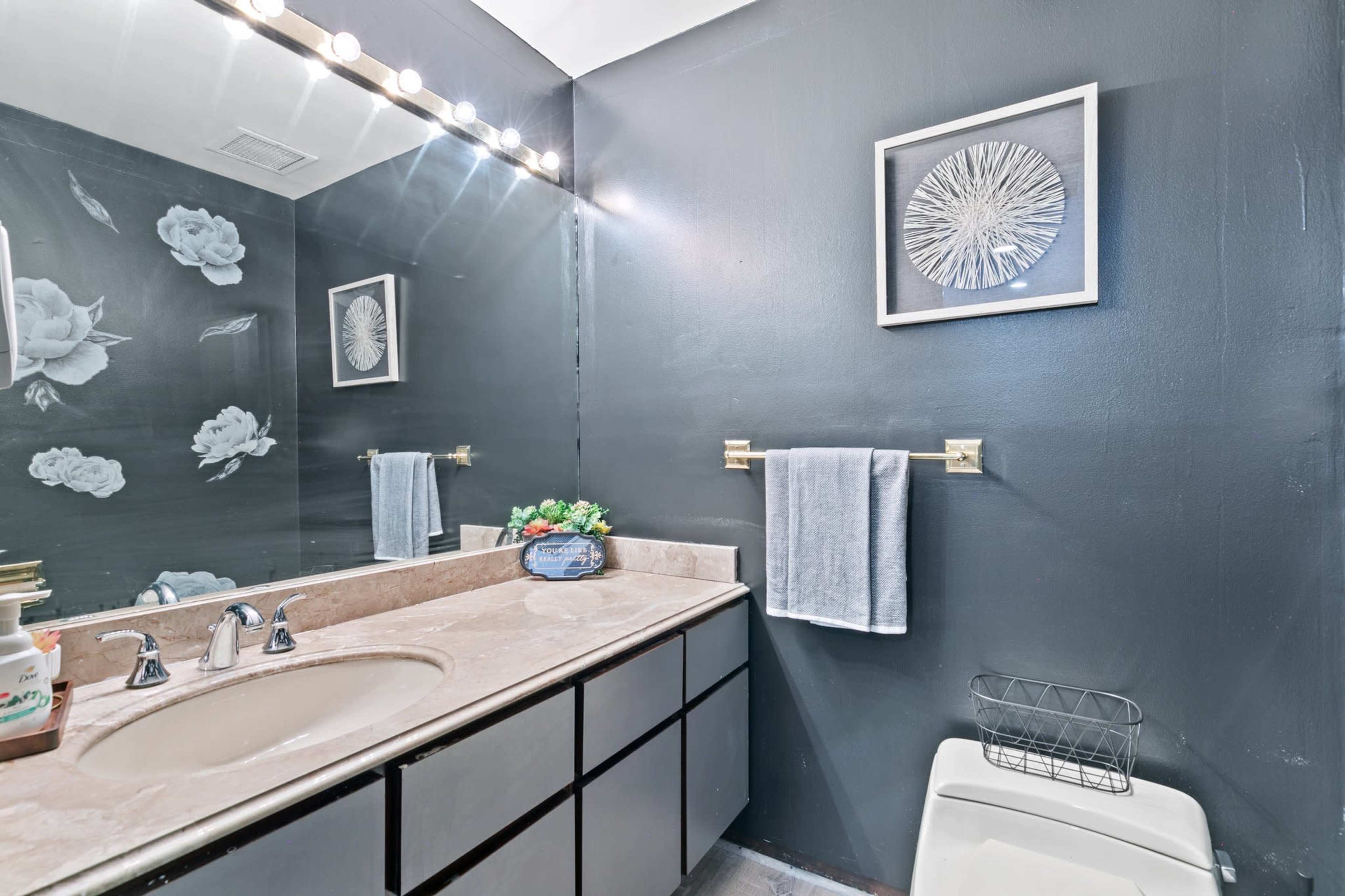 A bathroom with a dark gray wall, a vanity with a sink, a framed artwork, and a towel rack with towels.