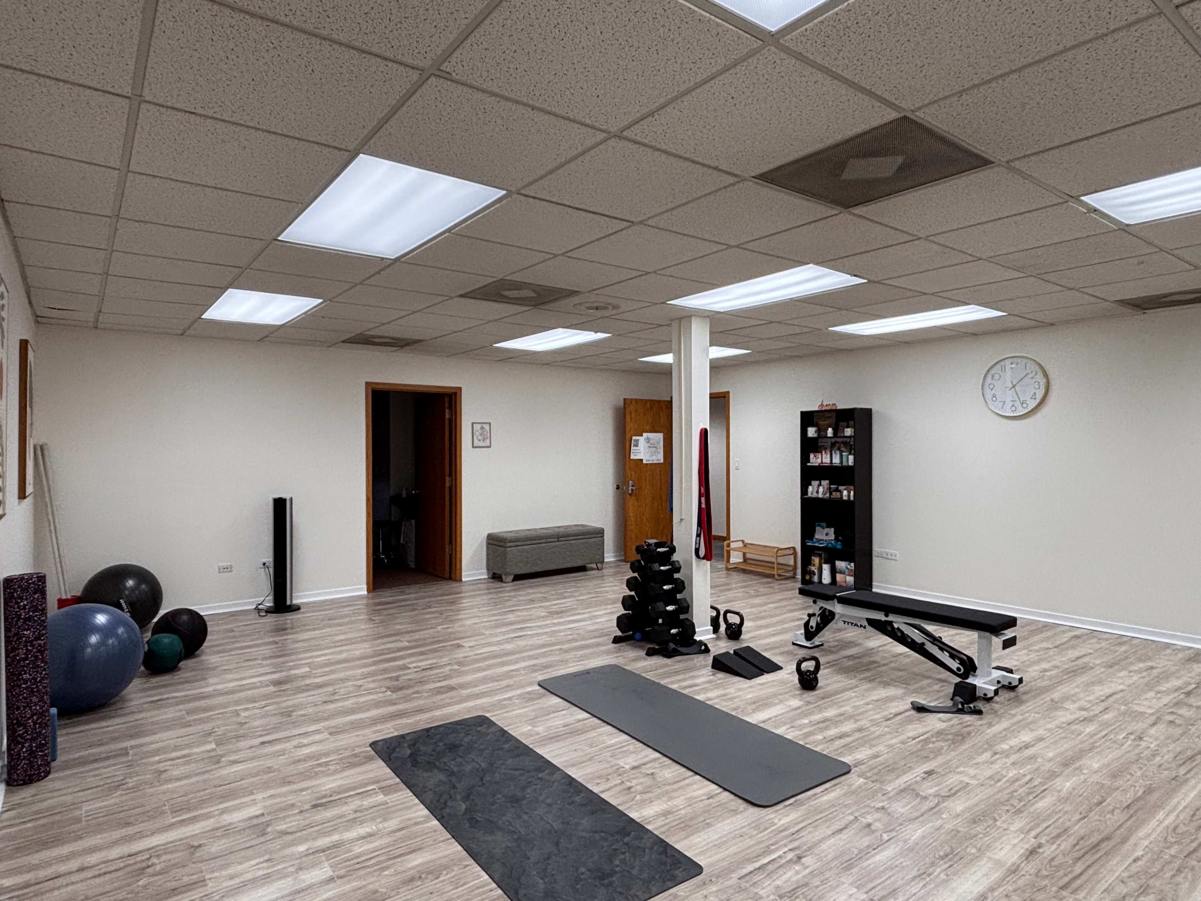 The image shows a spacious, well-lit exercise room with various fitness equipment, including weights, yoga mats, and a bench.