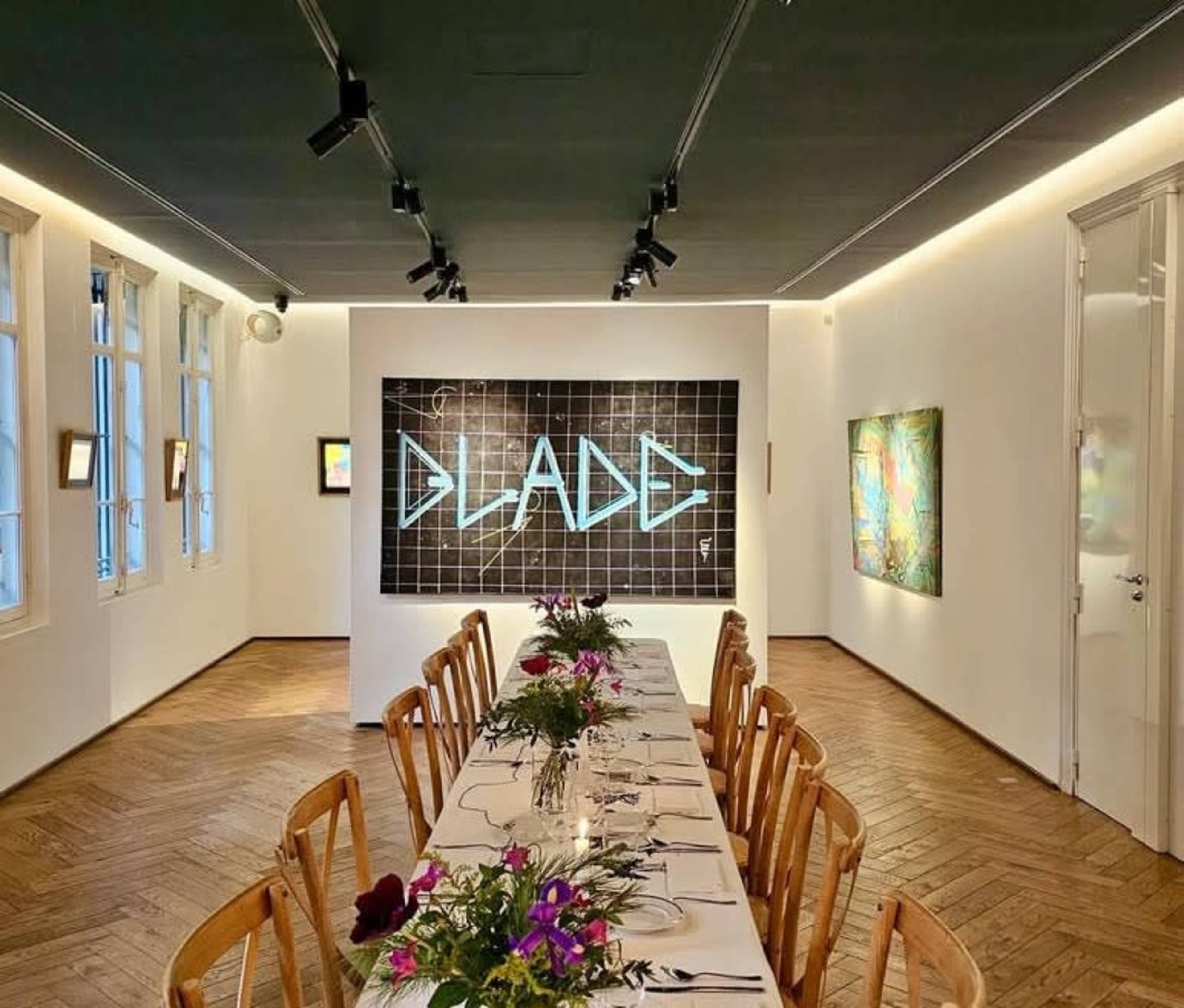 The image shows a dining room with a long table set for a meal, featuring a large neon artwork on the wall and artworks displayed around the room.
