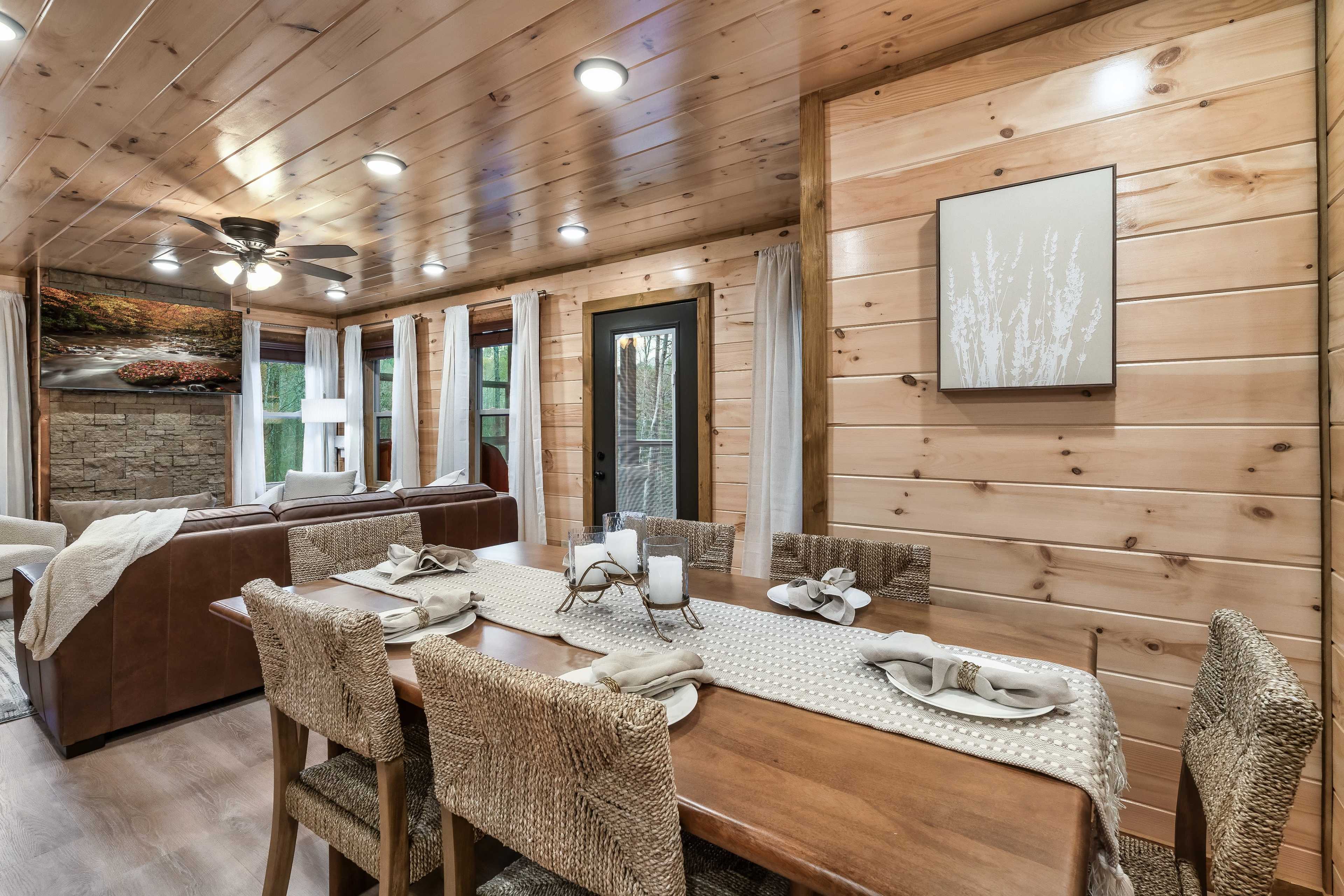 The image shows a cozy dining area with a wooden table set with decorative place settings, surrounded by a rustic interior featuring wooden walls and a large window.