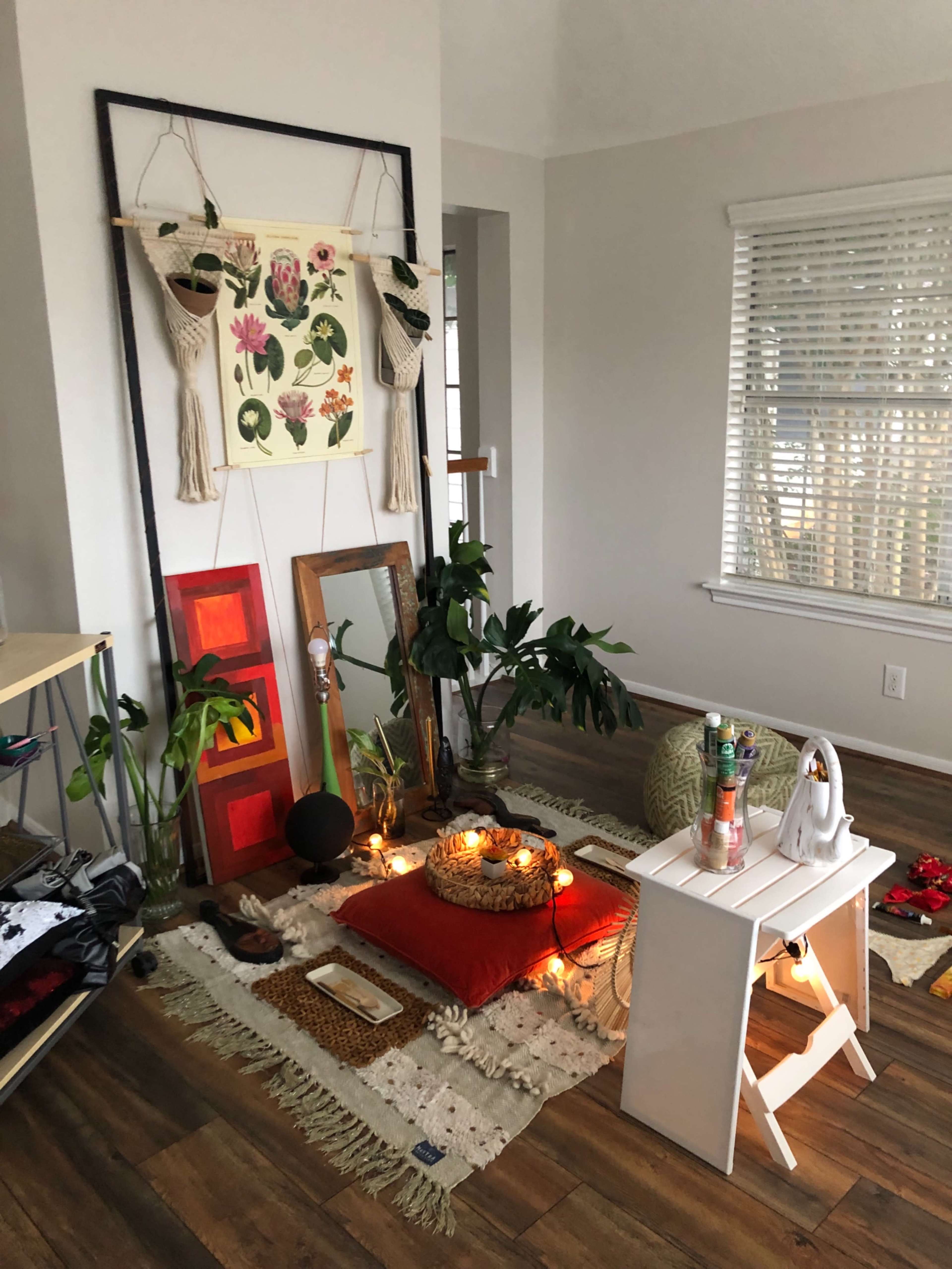 A cozy indoor space with a decorative wall hanging, several plants, a small white table, and various decorative elements on the floor.
