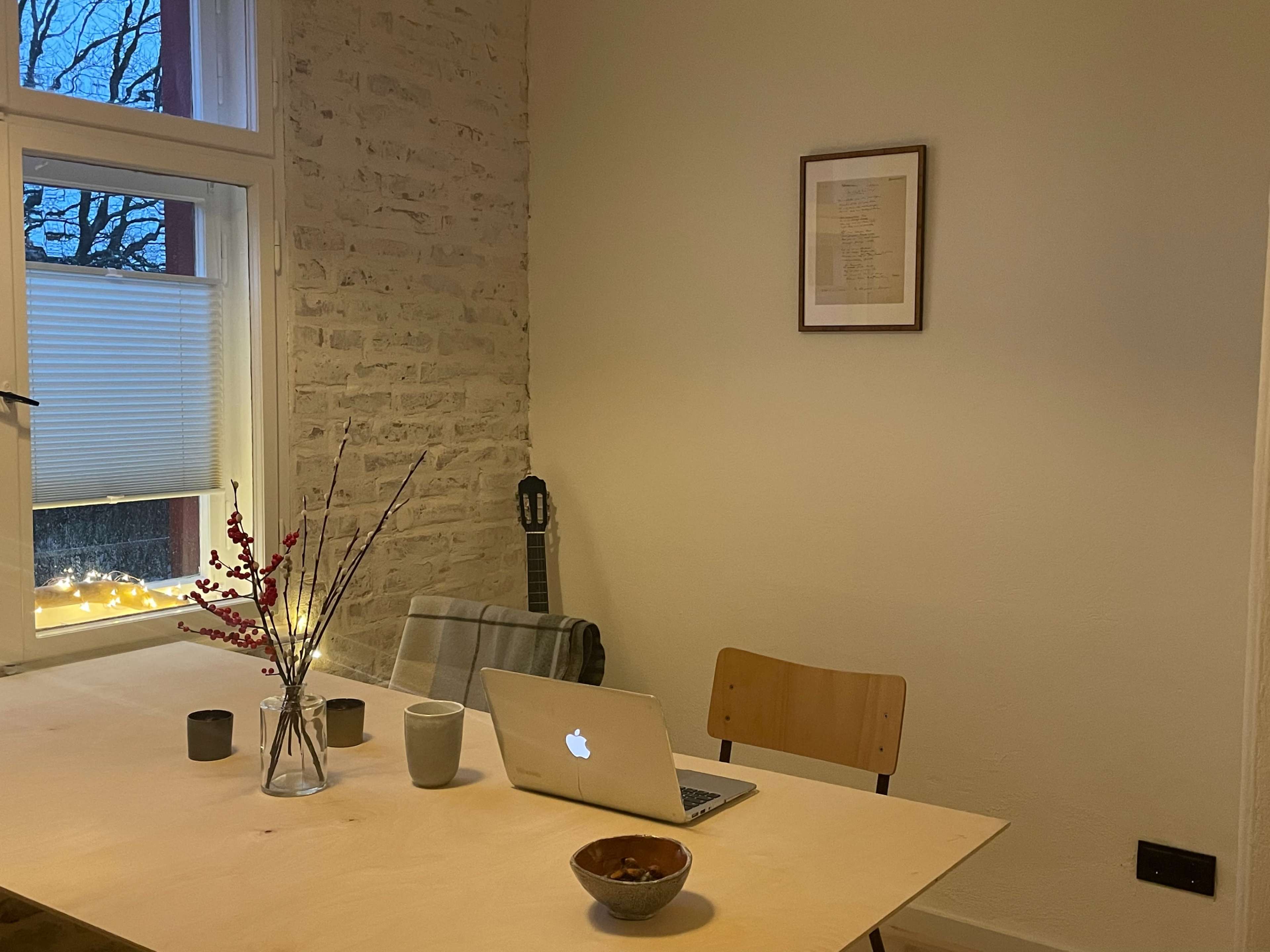 A simple dining table with a laptop, a bowl, two candles, and decorative branches, set against a light-colored wall with a framed picture and a guitar in the background.