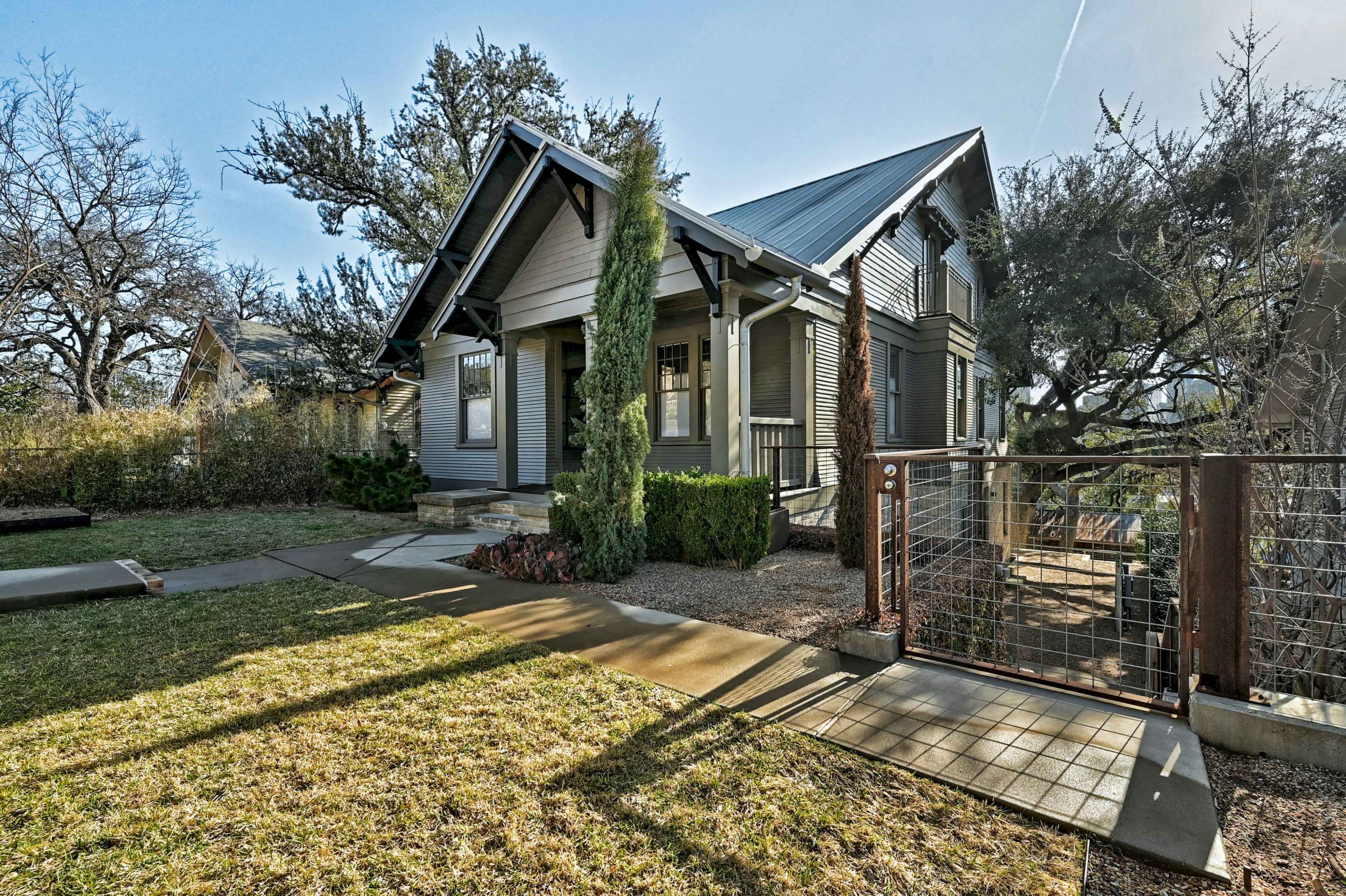 The image shows a two-story house with a sloped roof, surrounded by greenery and a pathway leading to a front gate.