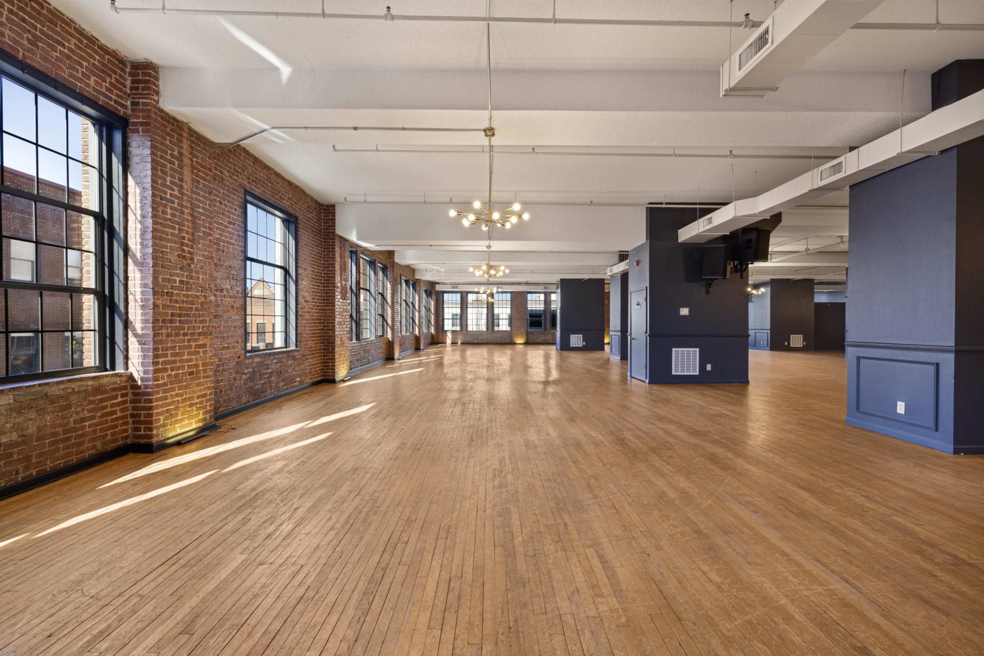 The image shows a spacious, well-lit room with large windows, exposed brick walls, and wooden flooring.