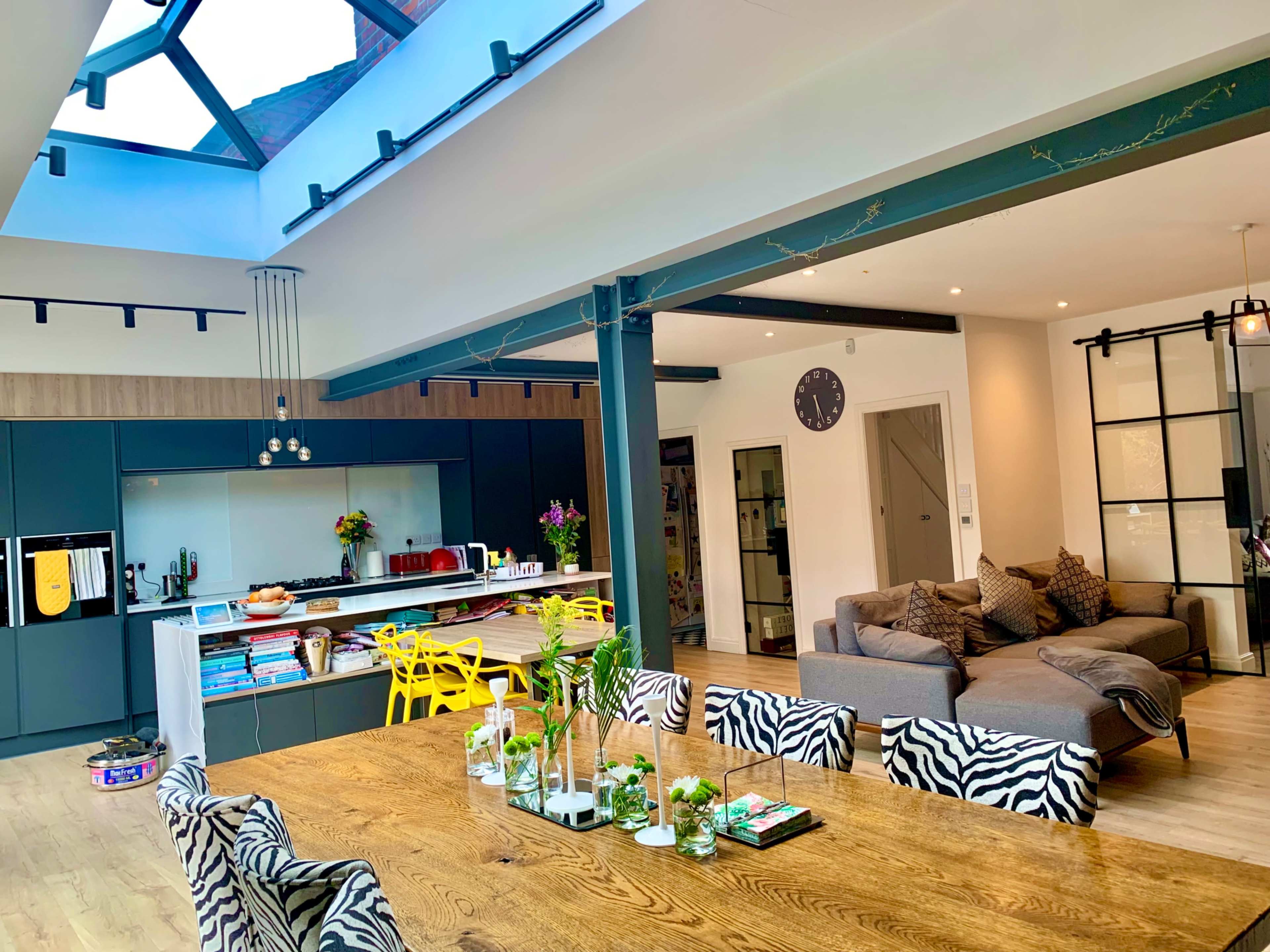 A modern kitchen and dining area with a large wooden table, vibrant yellow chairs, and a glass roof that allows natural light to enter the space.