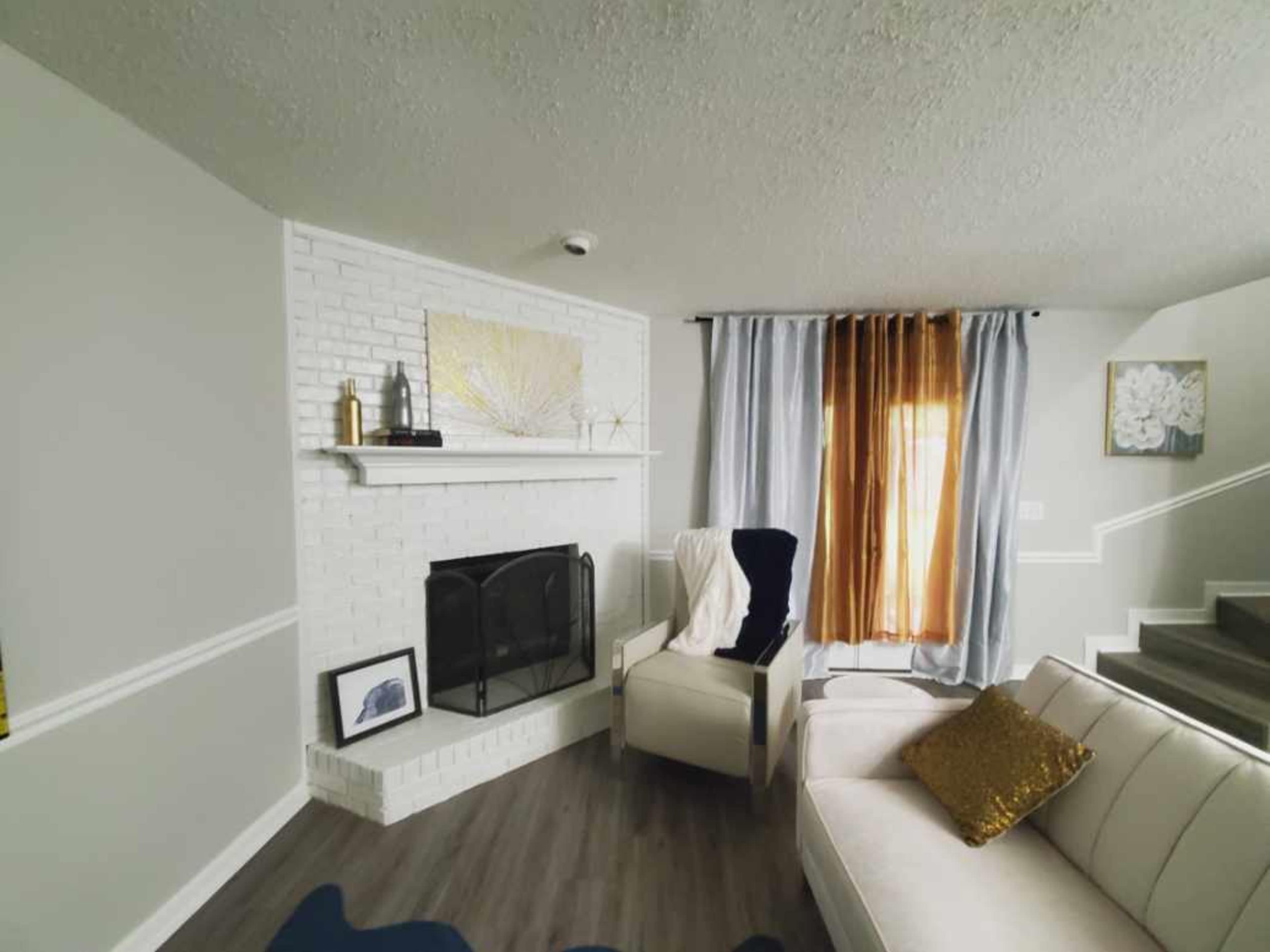 A living room features a white brick fireplace with a mantel, a light-colored sofa, and stairs leading to an upper level.