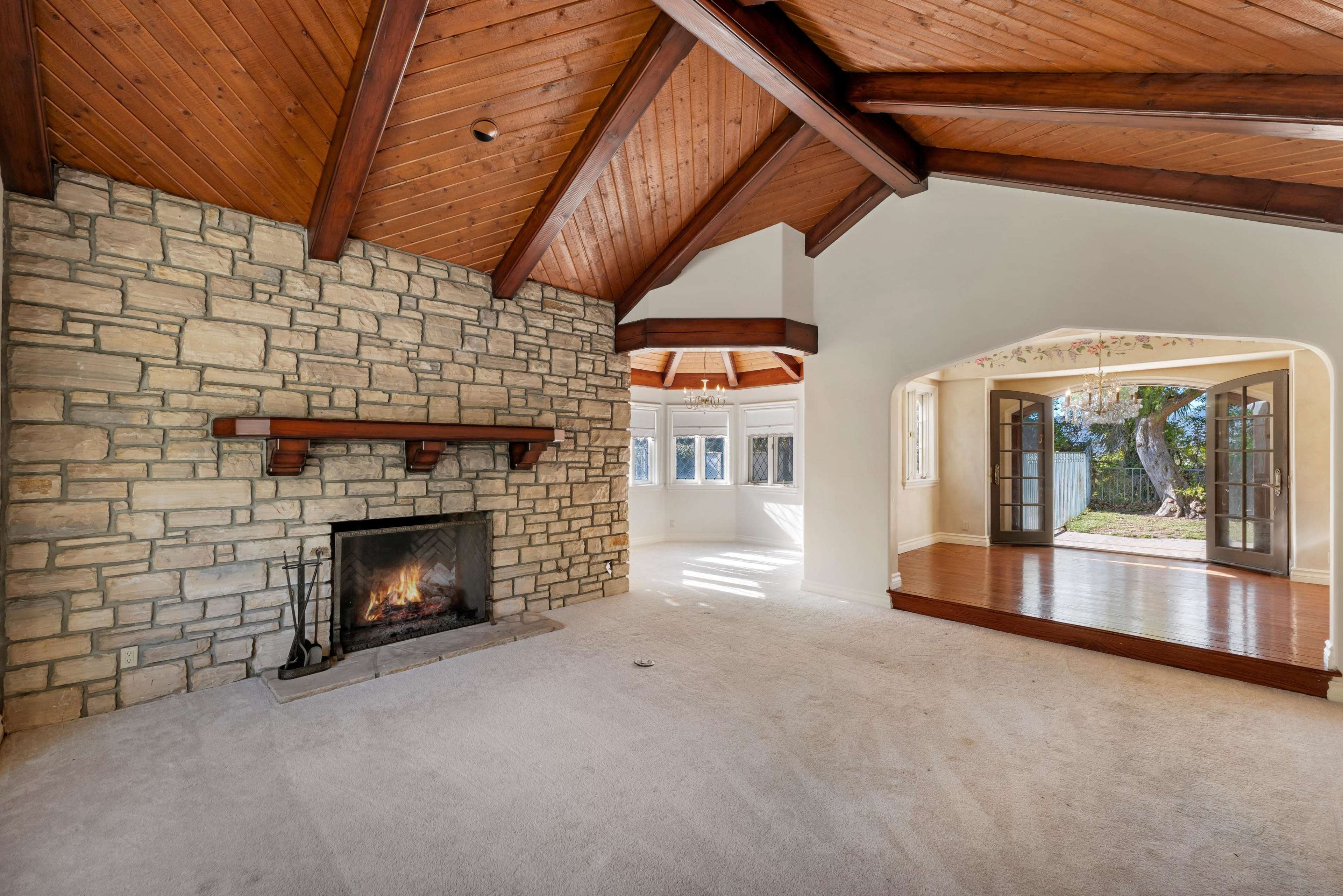 The image shows a spacious living room featuring a stone fireplace, wooden beams on the ceiling, and an adjoining room with double doors leading to an outdoor area.
