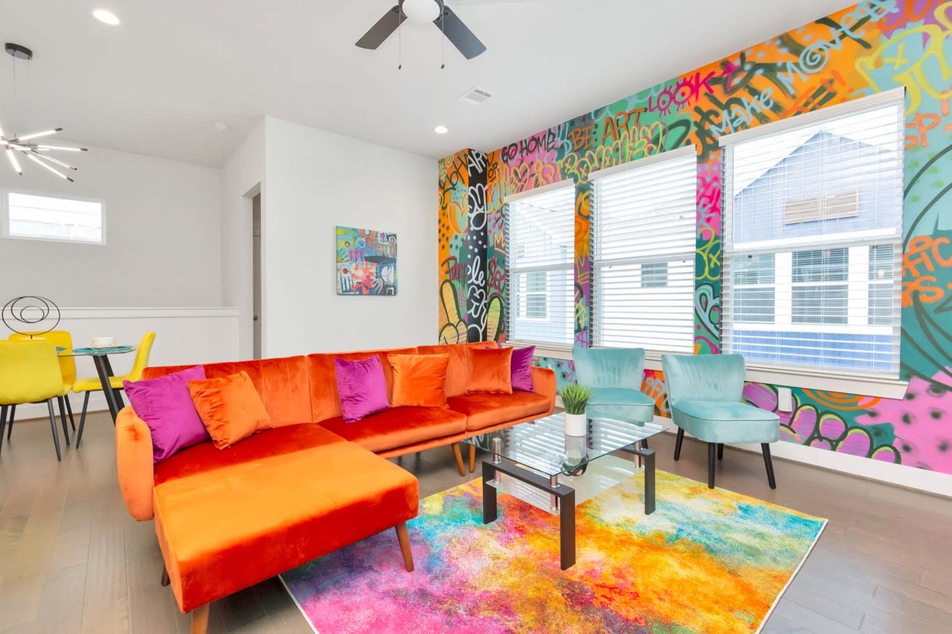 A vibrant living room features an orange and purple sectional couch, a colorful area rug, and a graffiti-style accent wall.