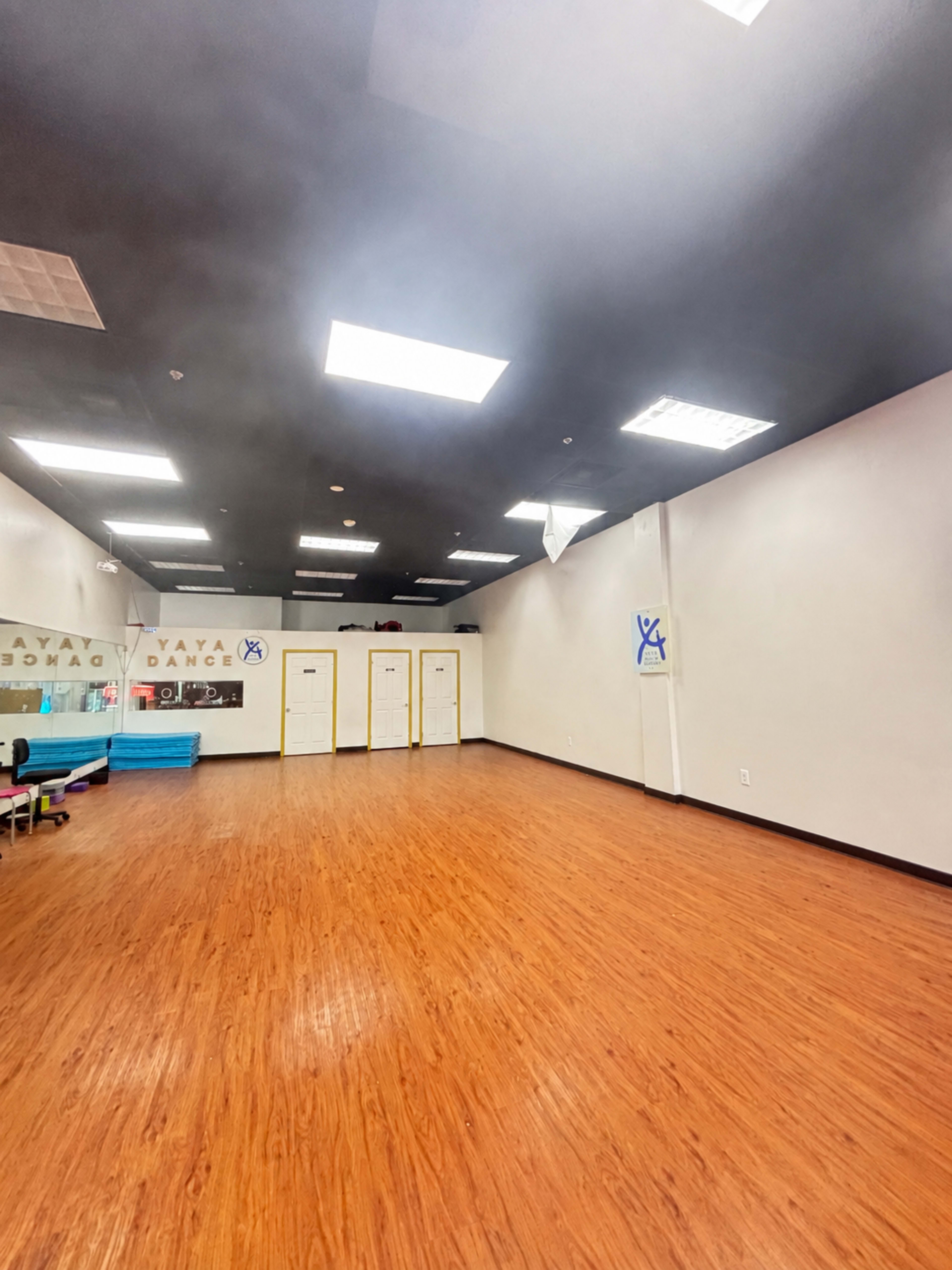 The image shows an empty dance studio with a wooden floor, white walls, and bright overhead lighting.
