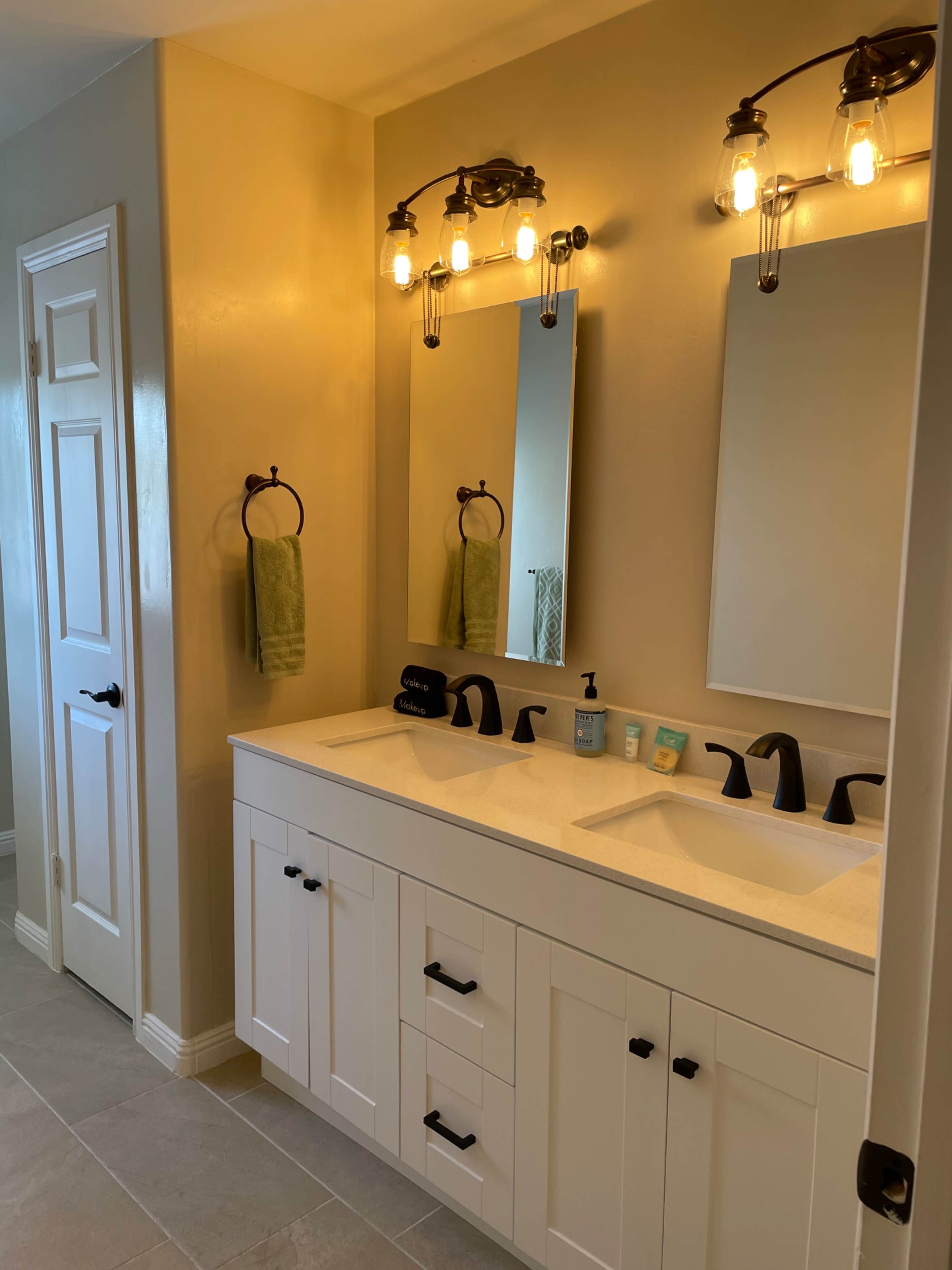 A bathroom with a double vanity featuring two sinks, large mirrors, and a light fixture above, against a light-colored wall.