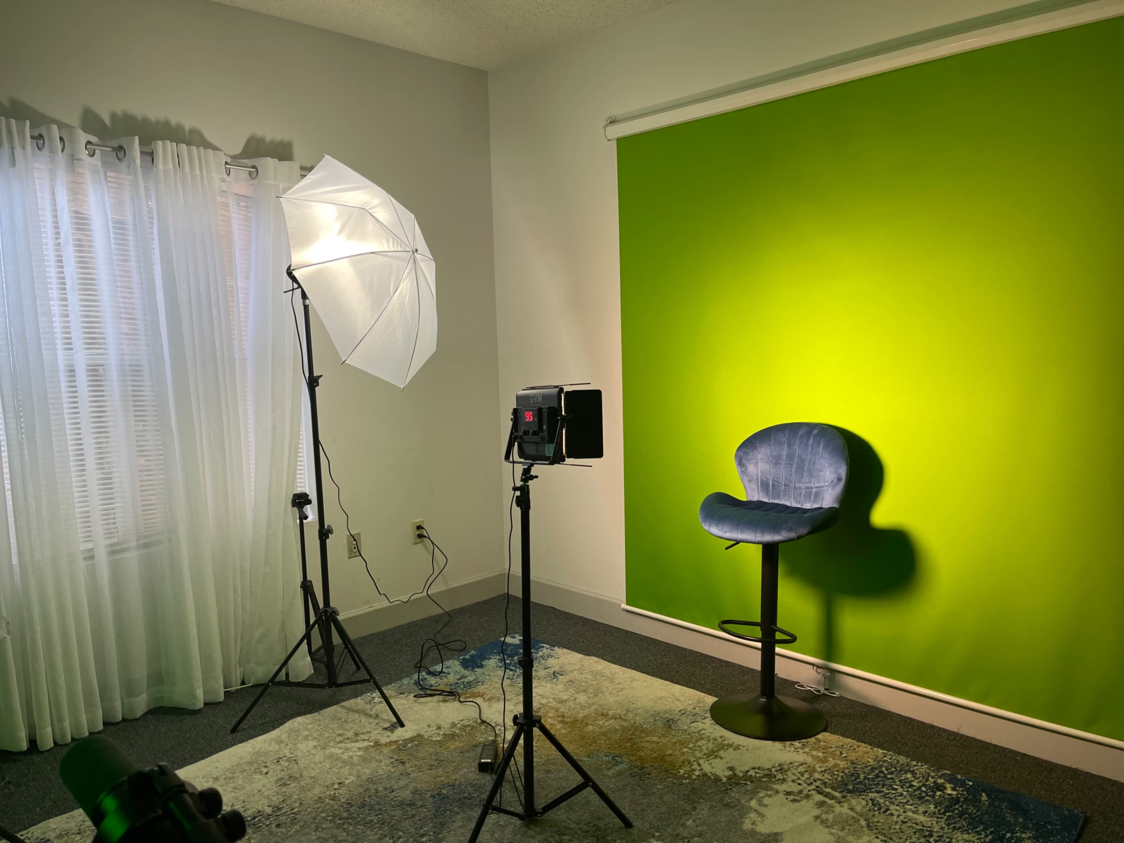 A minimal studio setup with a green backdrop, a blue chair, a lighting umbrella, and a spotlight aimed at the wall.