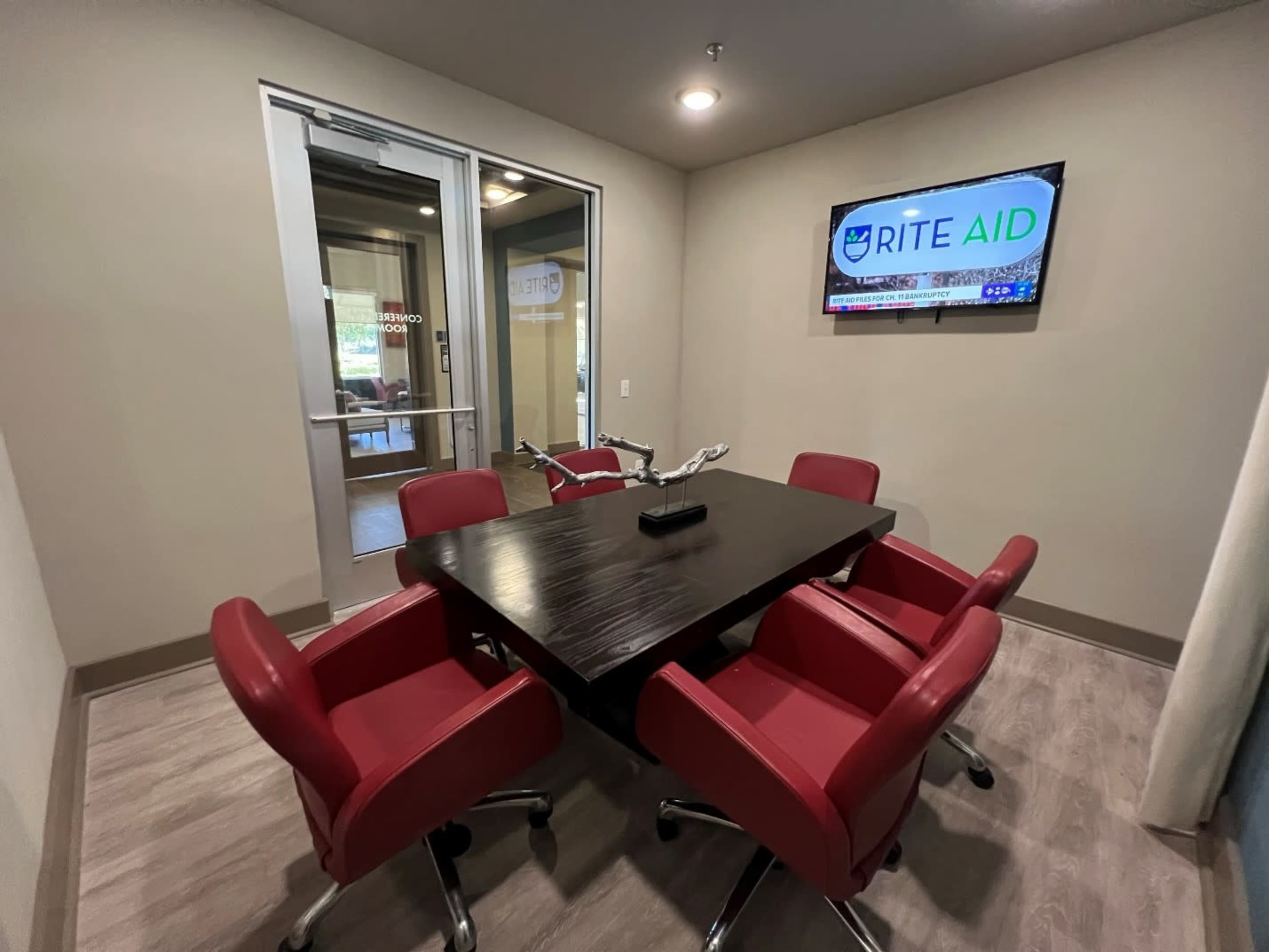 The image shows a small conference room with a dark wooden table surrounded by four red chairs and a television displaying the Rite Aid logo.