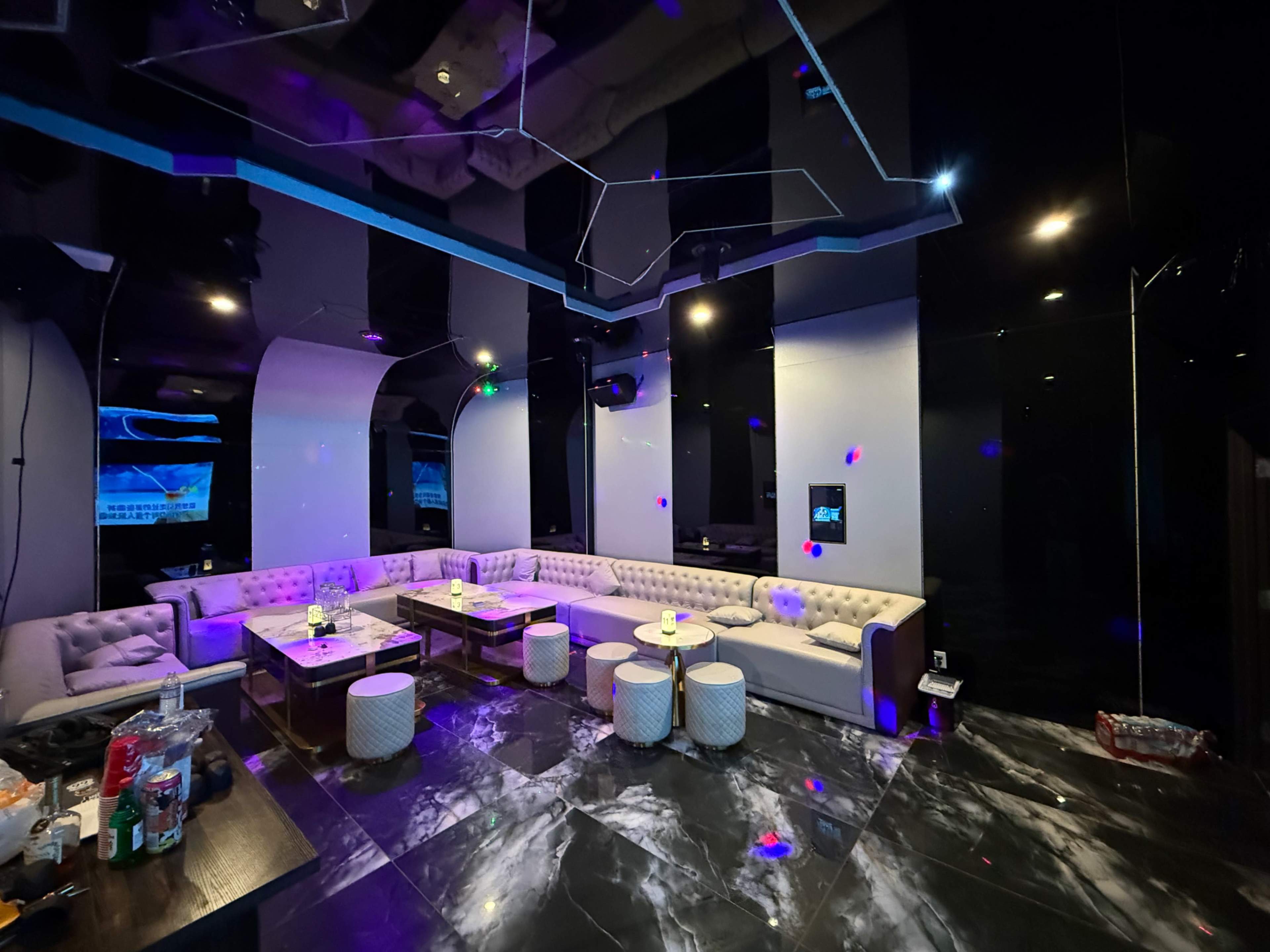 The image shows a modern, spacious lounge area with white sofas, small tables, and a sleek black and marble floor, illuminated by colorful lights.