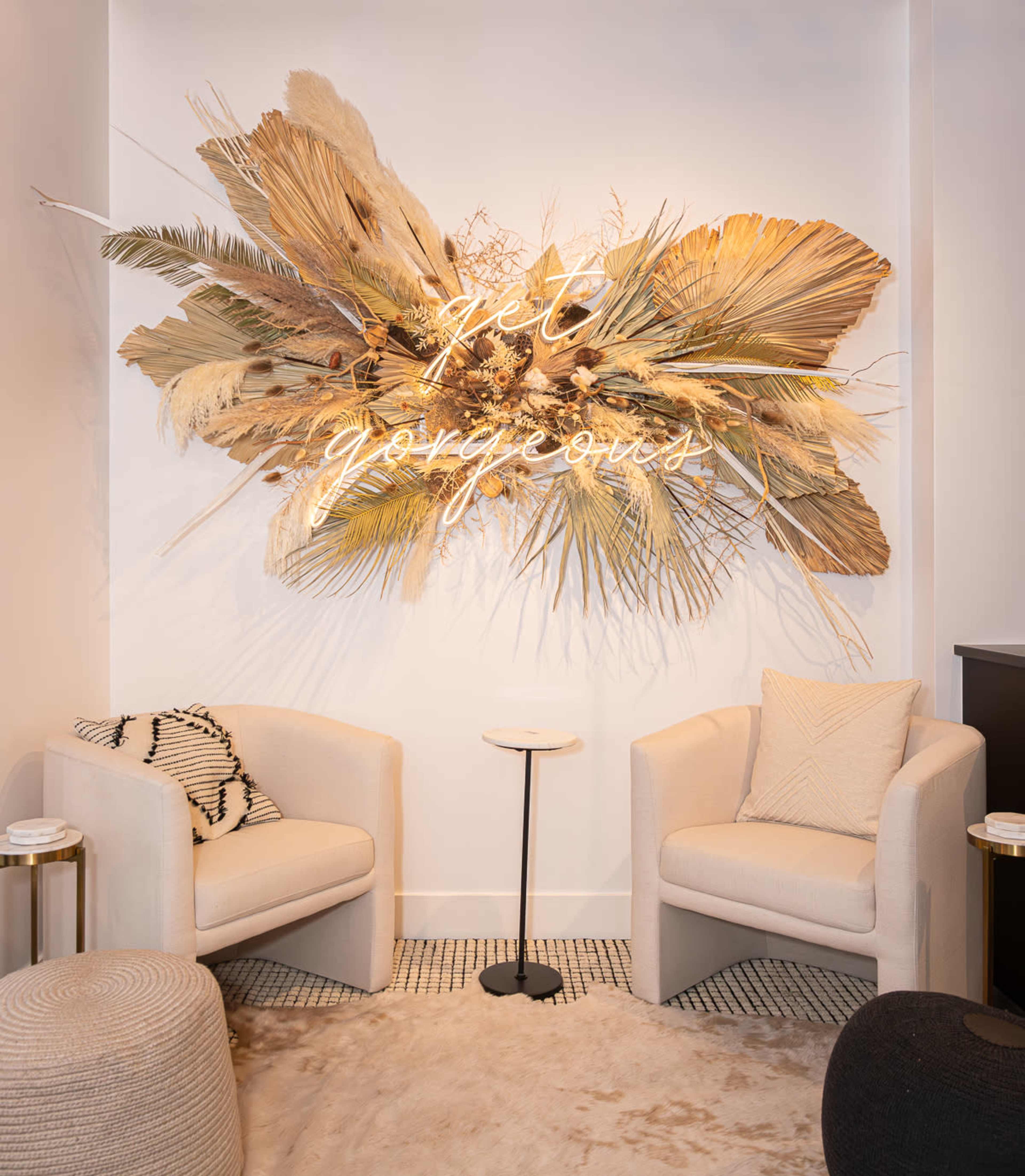 The image features a stylish seating area with two cream-colored armchairs facing a decorative wall adorned with dried plants and a neon sign that reads "Be Gorgeous."