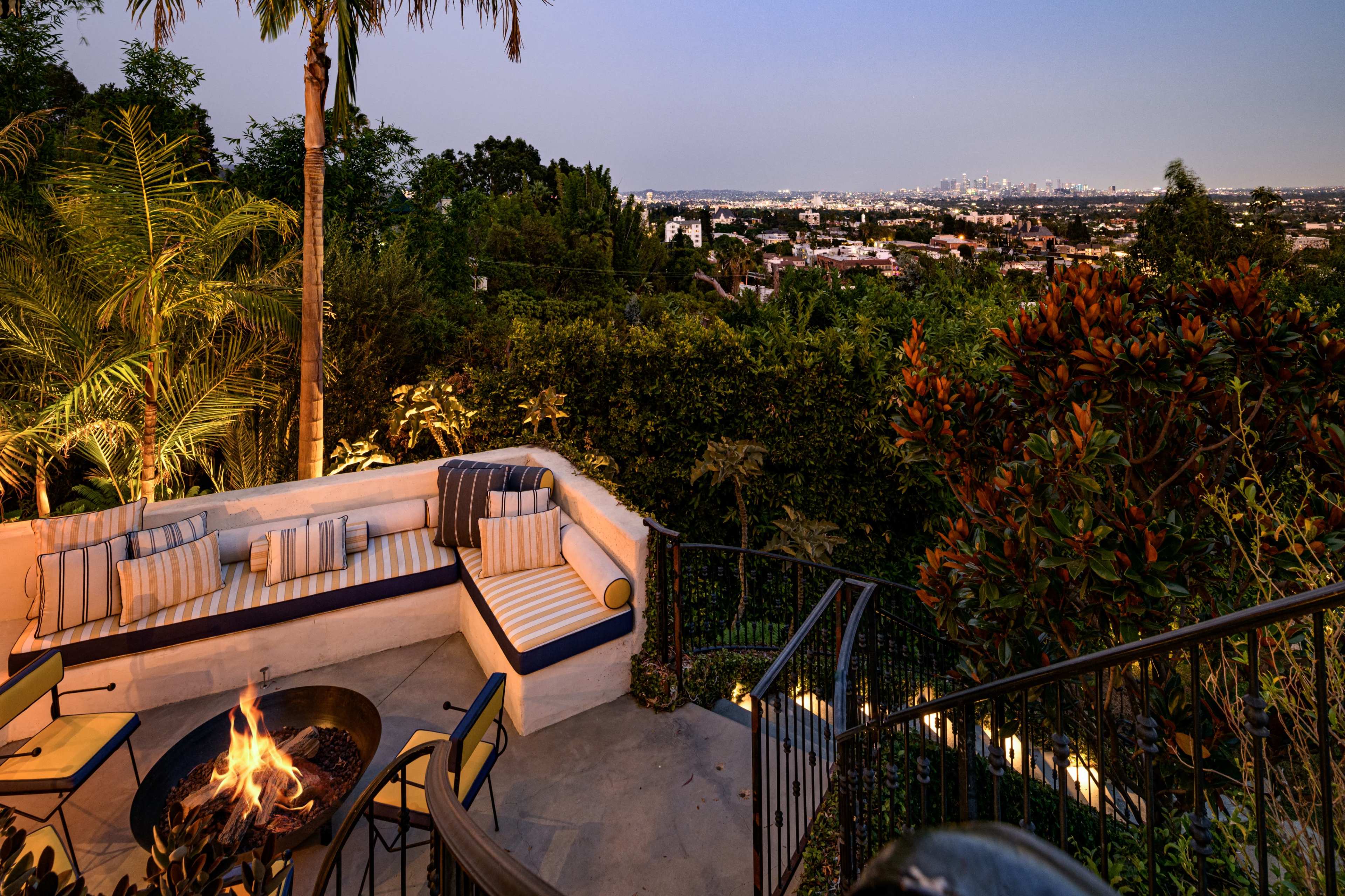 A hillside patio features a circular fire pit surrounded by seating, with city lights visible in the distance.