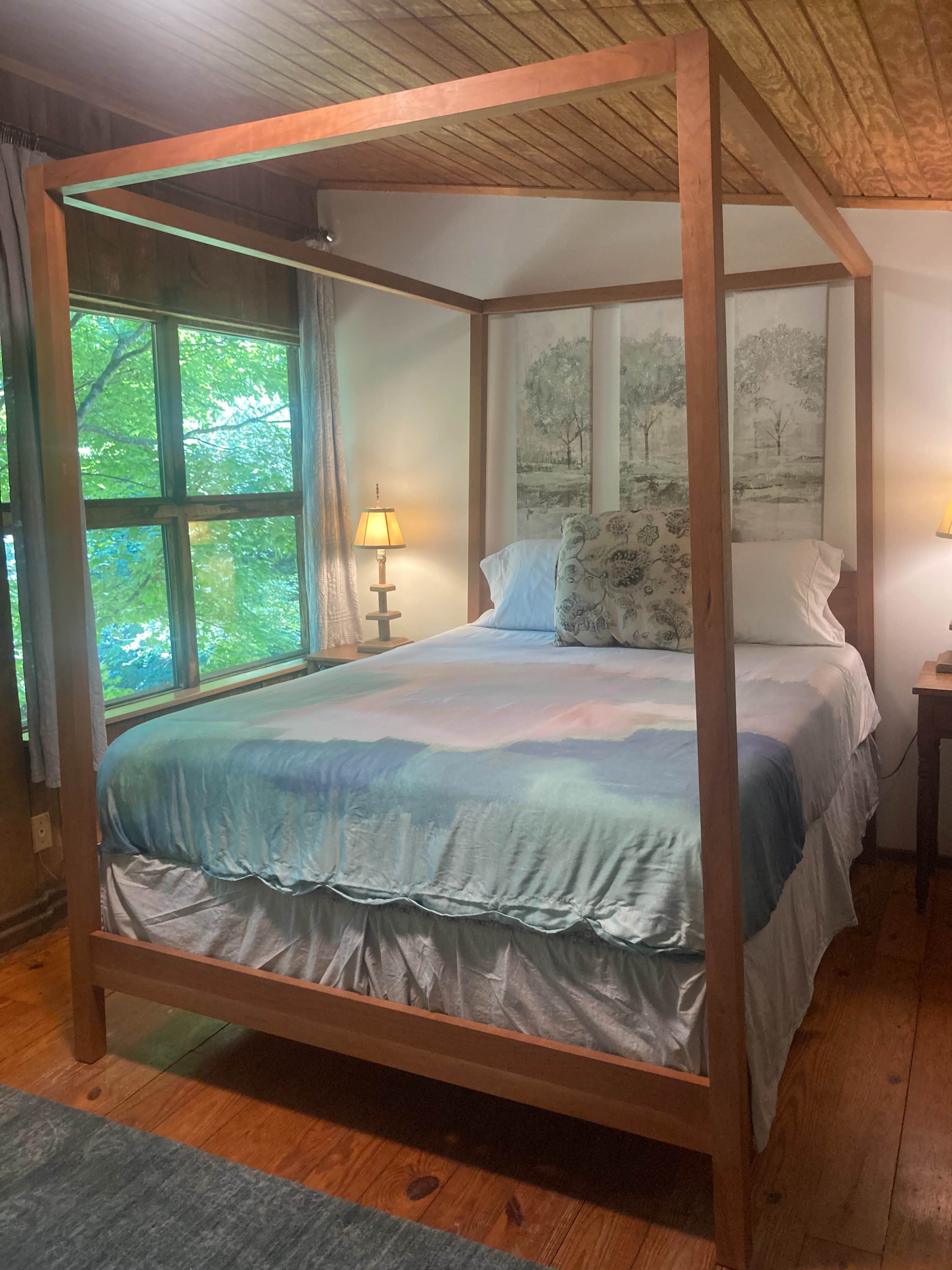 A bedroom with a four-poster bed, two lamps on nightstands, and large windows displaying greenery outside.