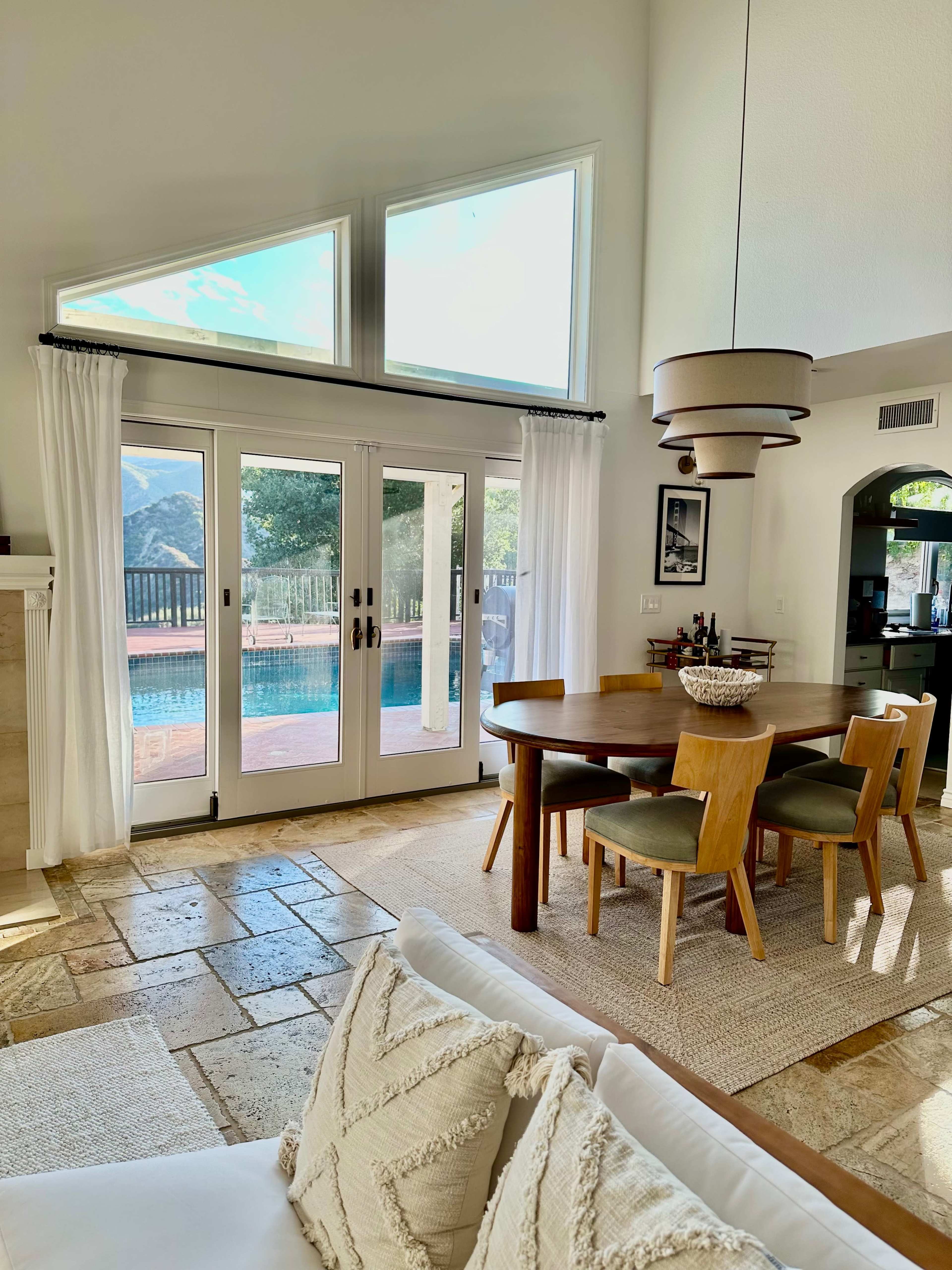 A dining area with a round wooden table and green chairs overlooks a patio with a swimming pool through large glass doors.