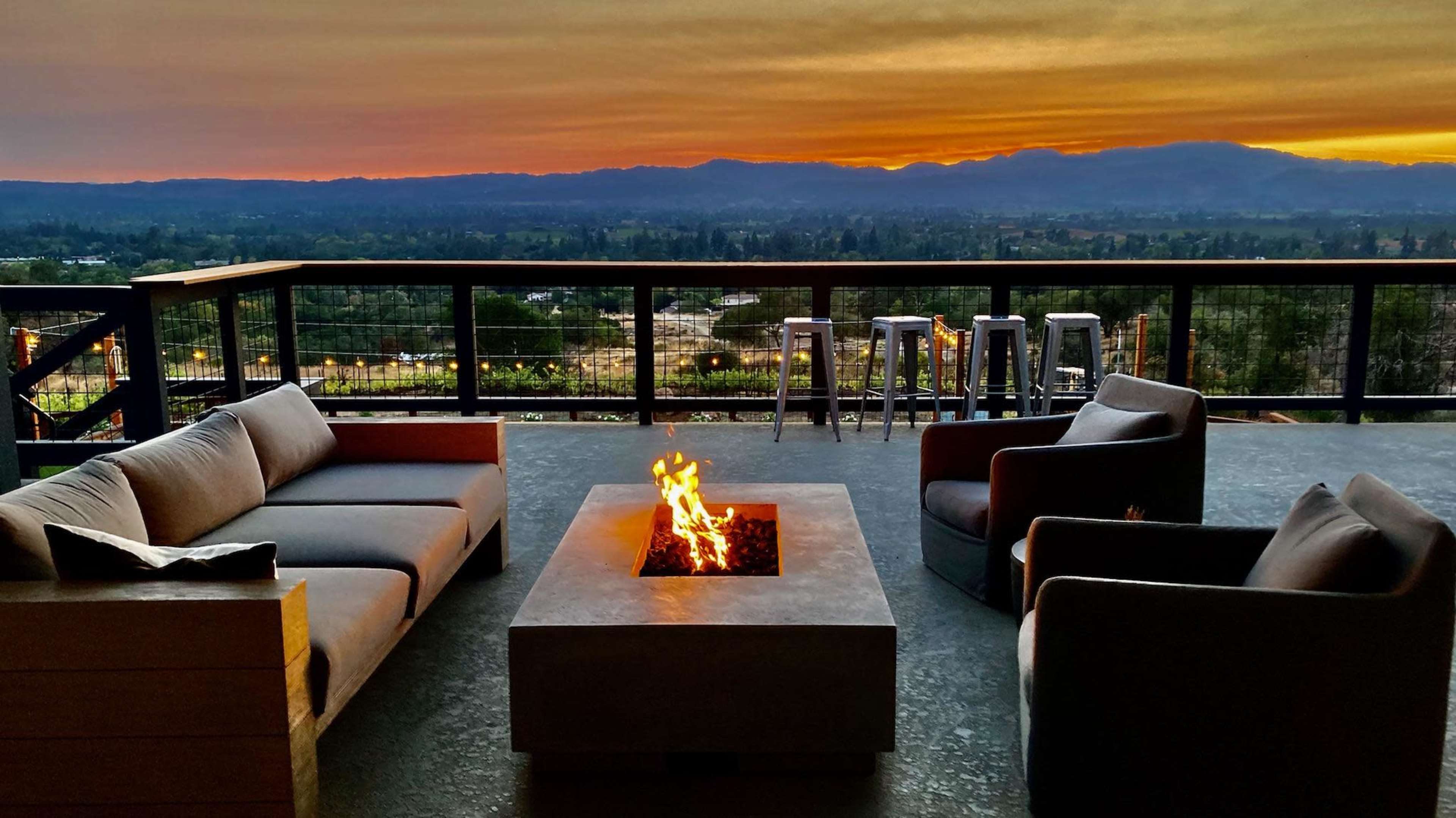 A patio area features modern seating around a central fire pit with a view of the sunset over distant mountains.