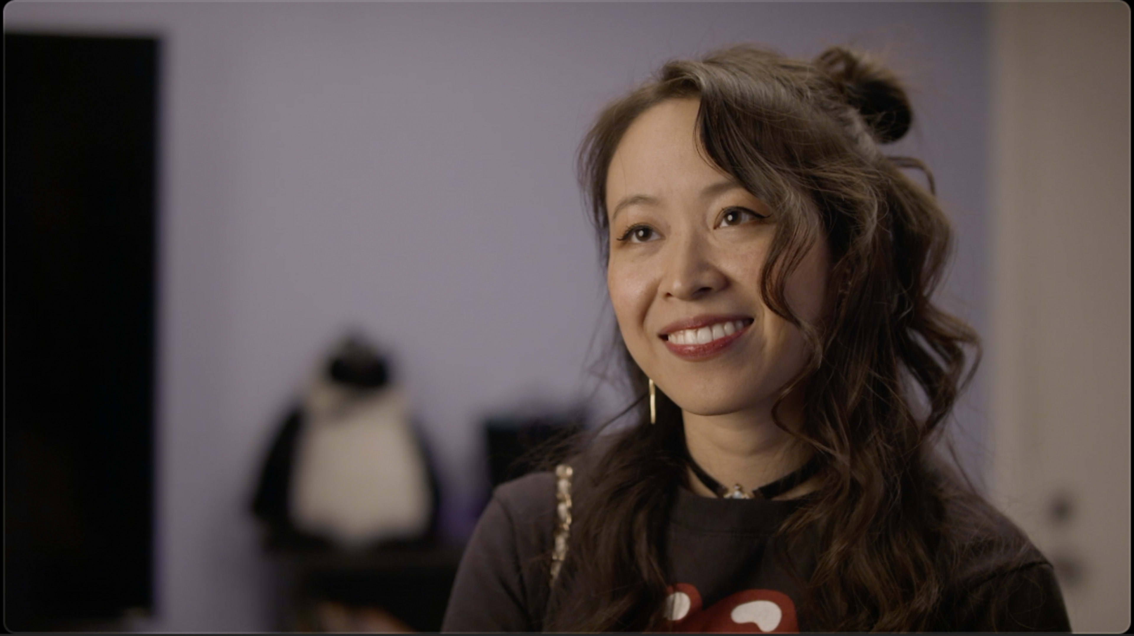 A woman with long, wavy hair smiles while sitting in a room with a purple wall and a plush toy in the background.