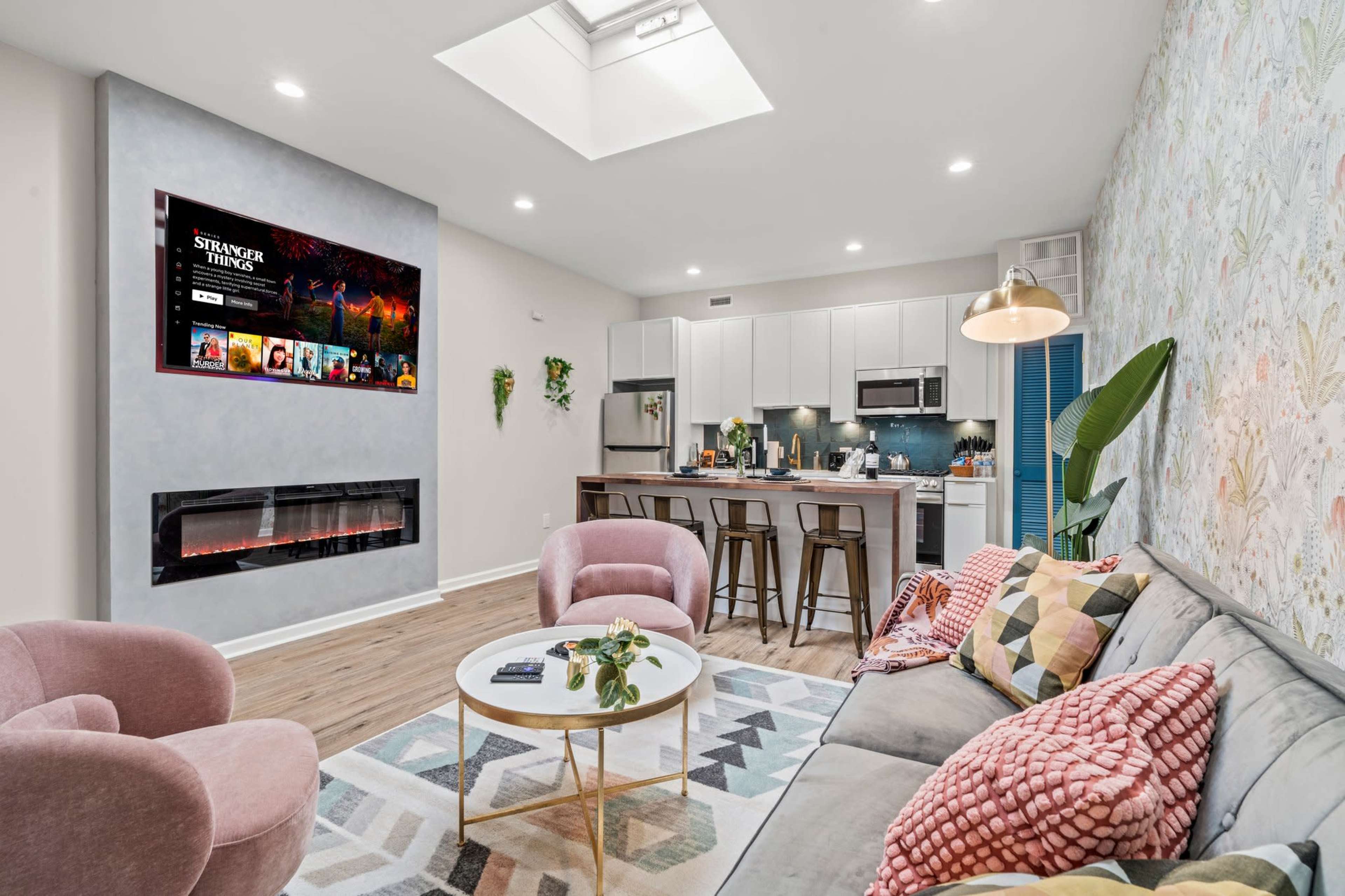 The image depicts a modern living room featuring a sofa with patterned cushions, a circular coffee table, a wall-mounted television showing a streaming interface, and a kitchen area in the background.