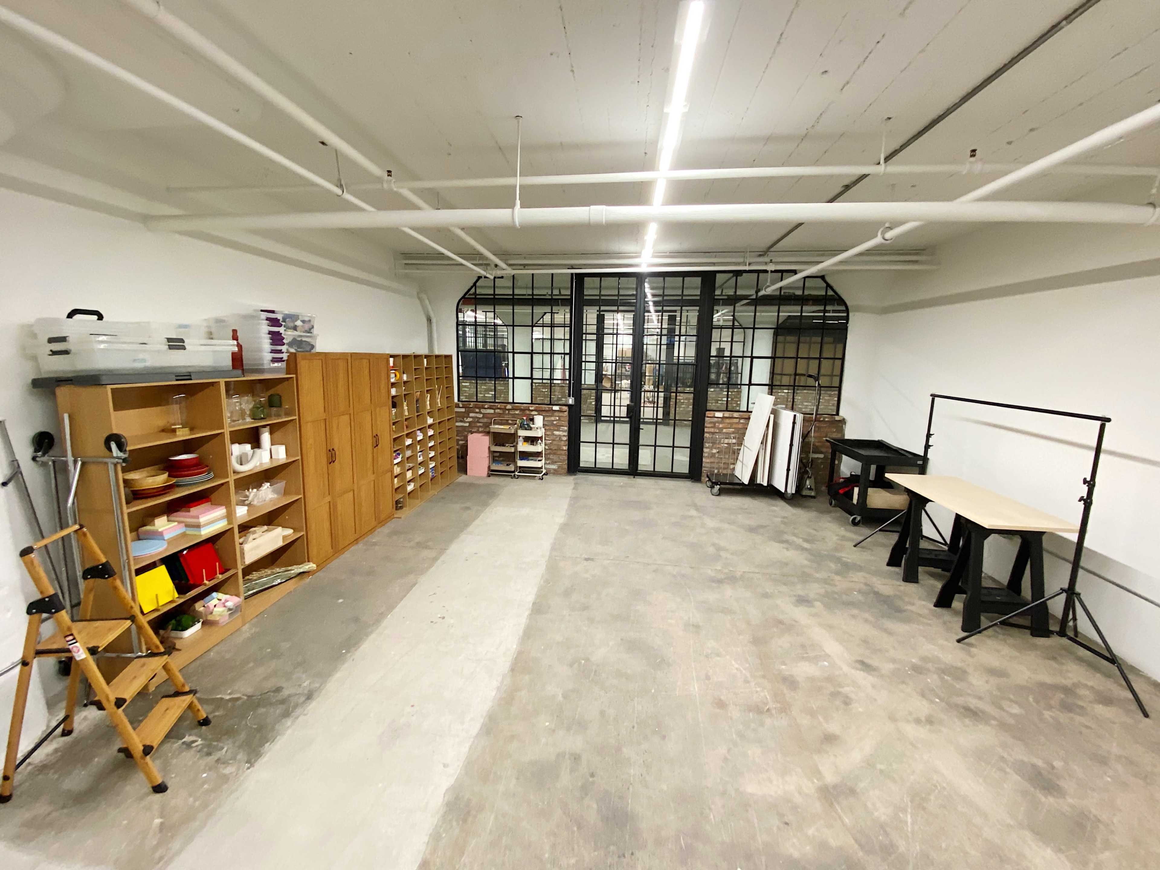 A spacious, empty room with a wooden shelving unit on the left, a table on the right, and large glass windows at the far end.