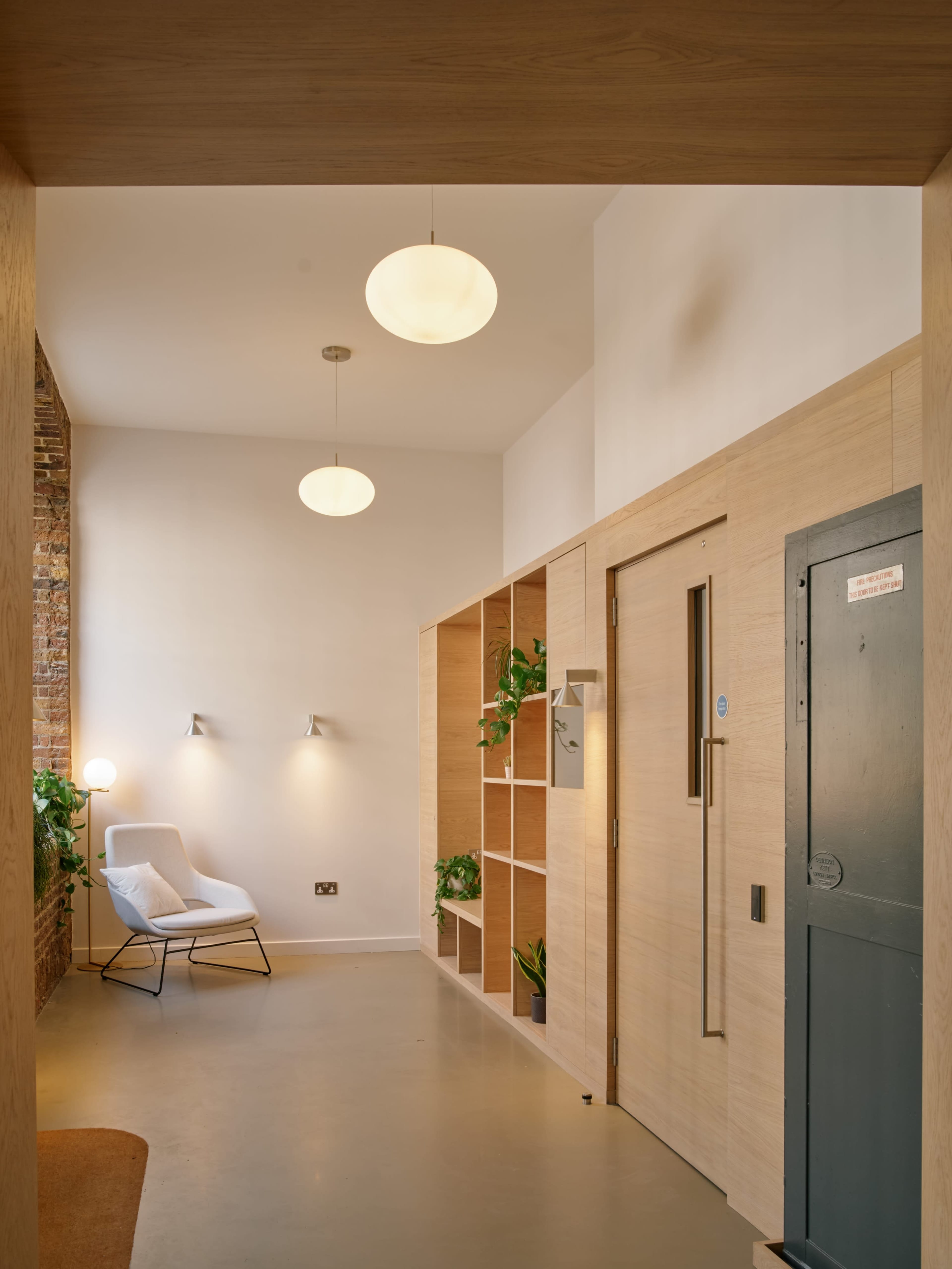 The image shows a modern entryway featuring a minimalist design with wooden shelving, a lounge chair, and plants, illuminated by pendant lights.