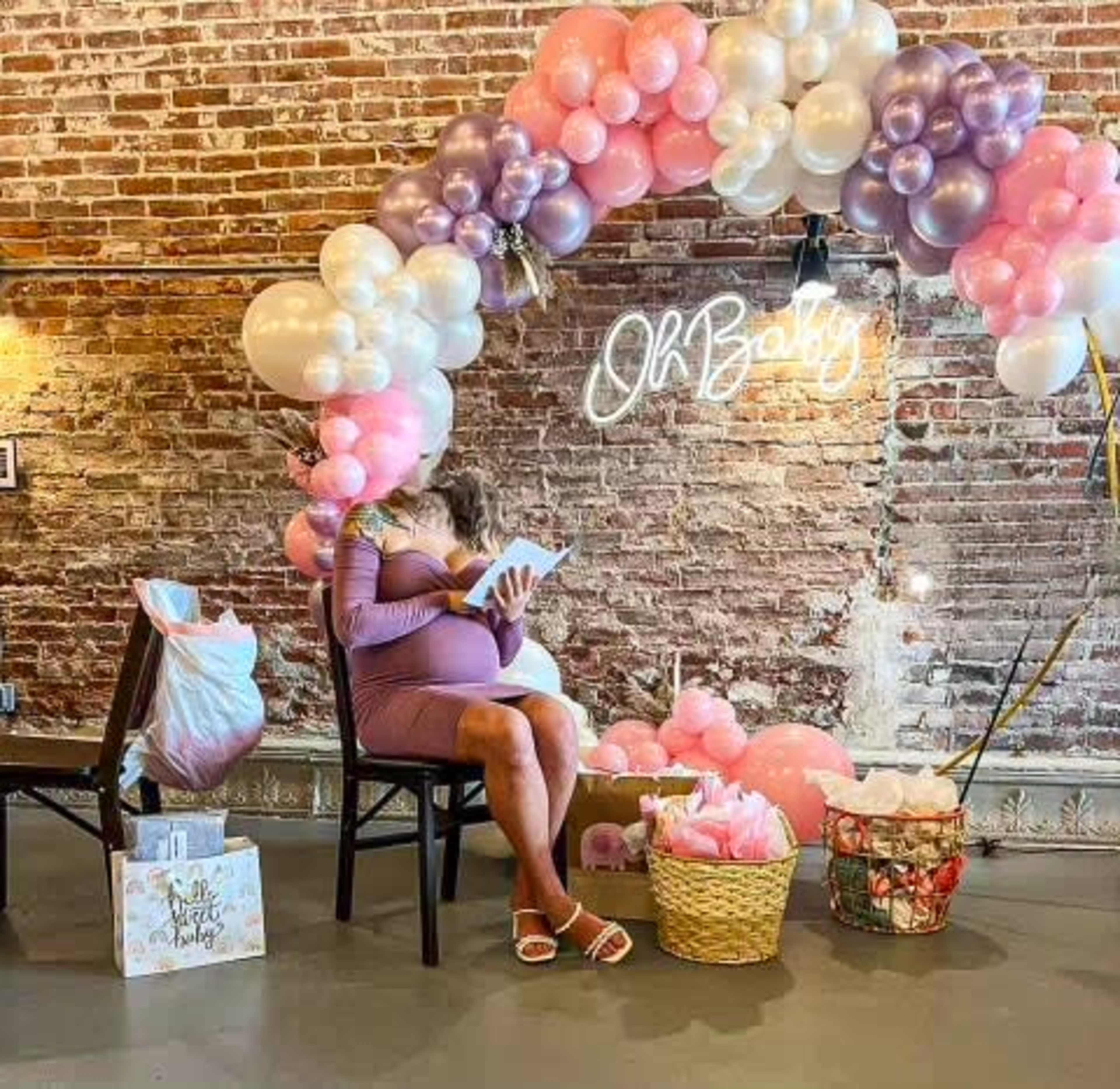 A pregnant woman sits on a chair reading a book under a balloon arch that says "Oh Baby."