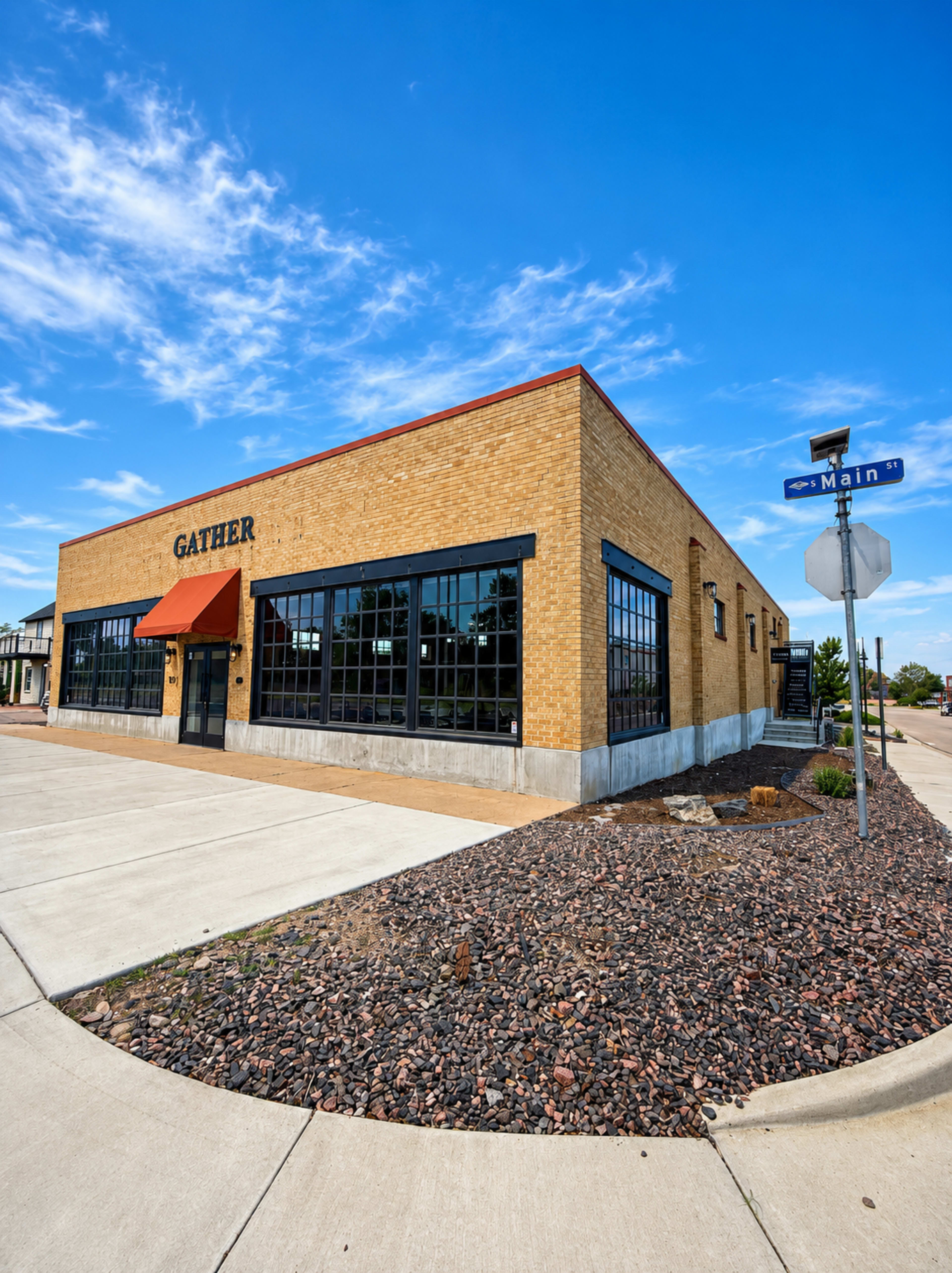 A brick building with large windows features the word "GATHER" on its facade, located at the intersection of Main Street and another road.
