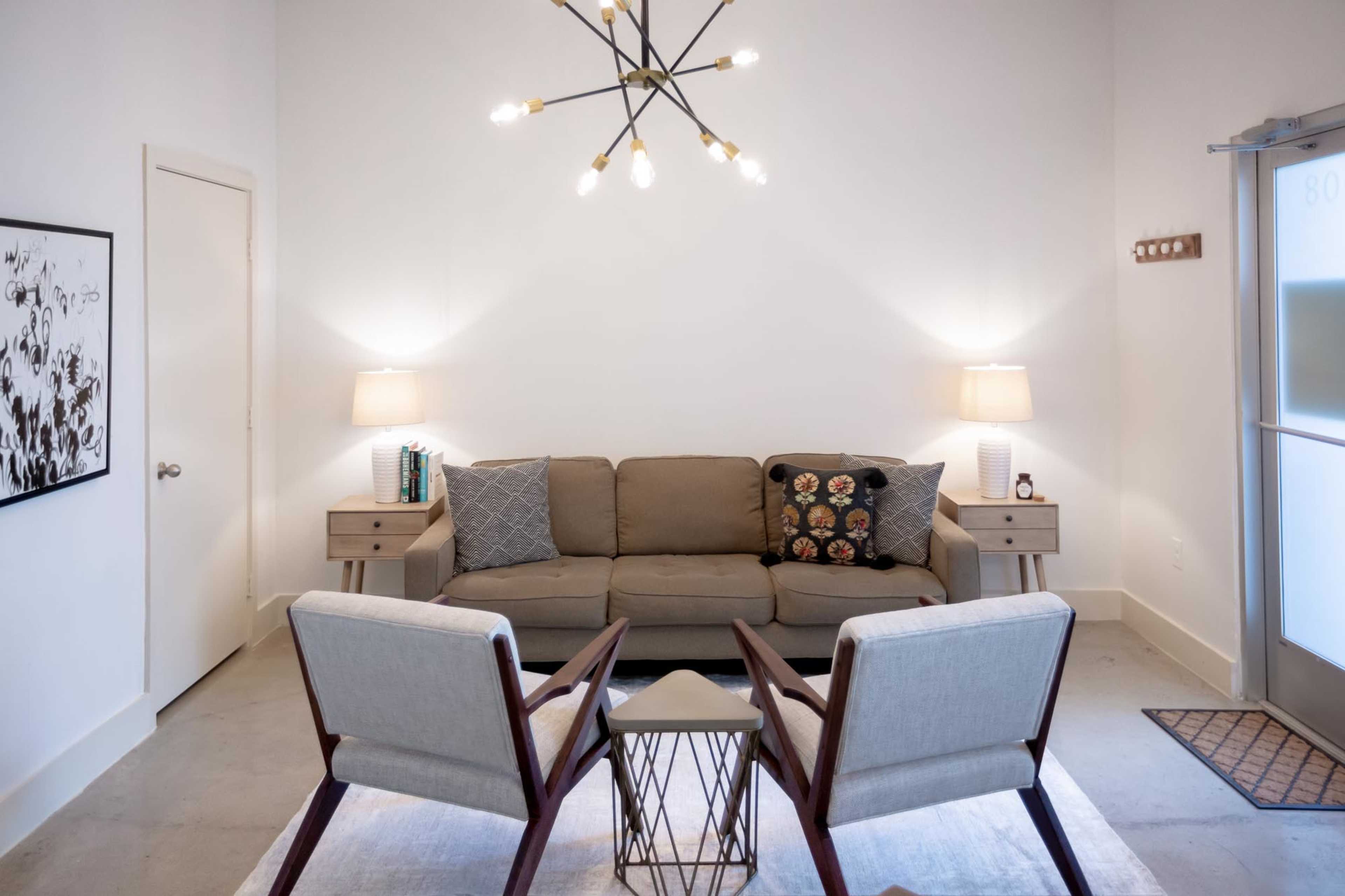 A minimalist living room with a beige sofa, two armchairs, and a coffee table under a modern chandelier.