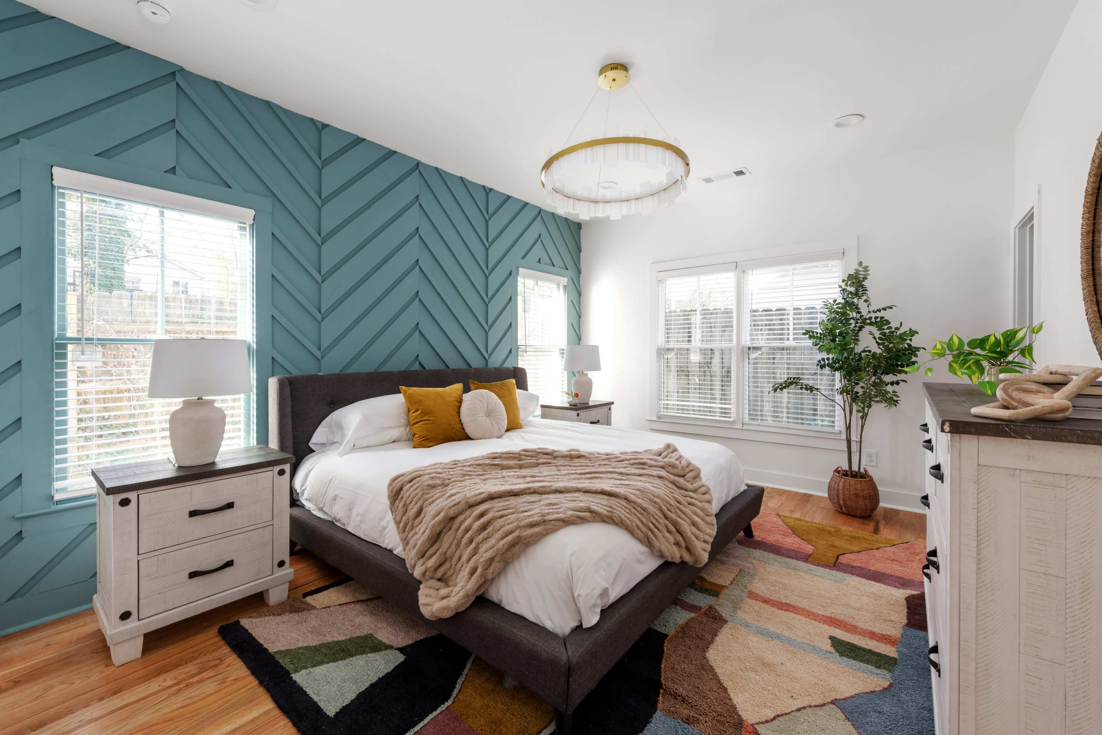 A modern bedroom features a teal herringbone accent wall, a gray bed with decorative pillows, and a colorful area rug.