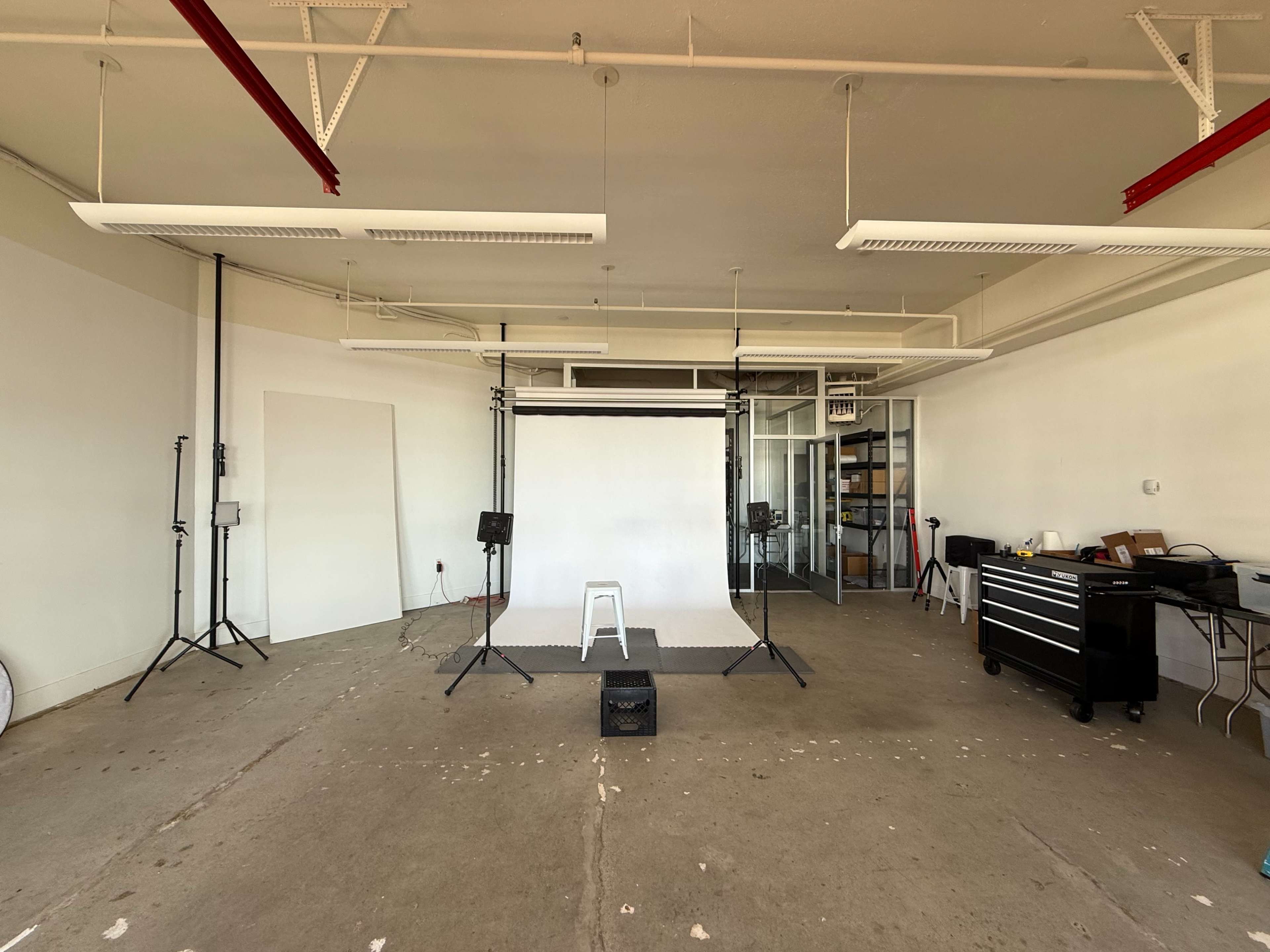 The image shows a spacious studio setup with a white backdrop, two light stands, a stool, and storage shelving in the background.