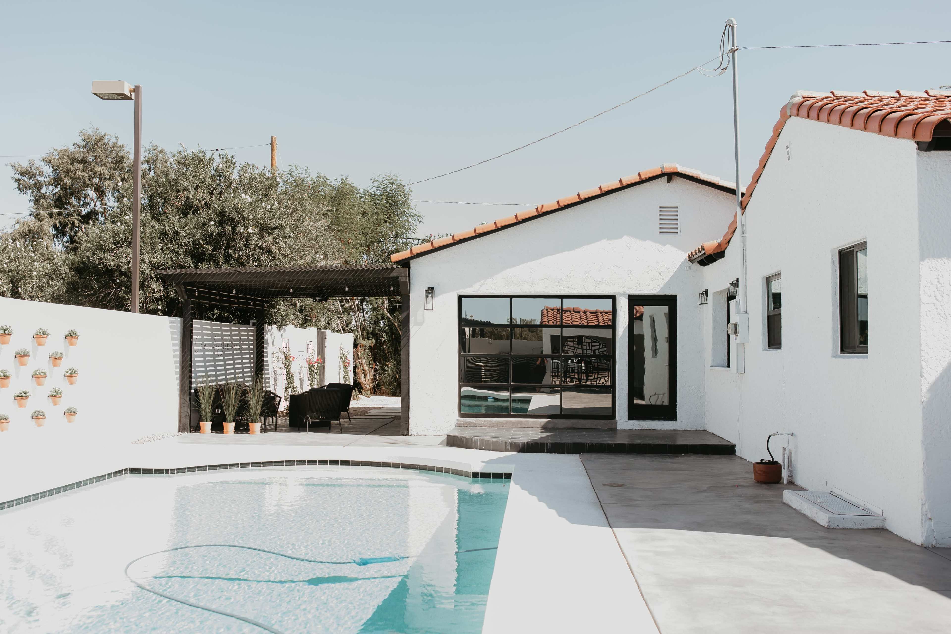 A modern white house with a tiled roof overlooks a swimming pool surrounded by plants and outdoor furniture.