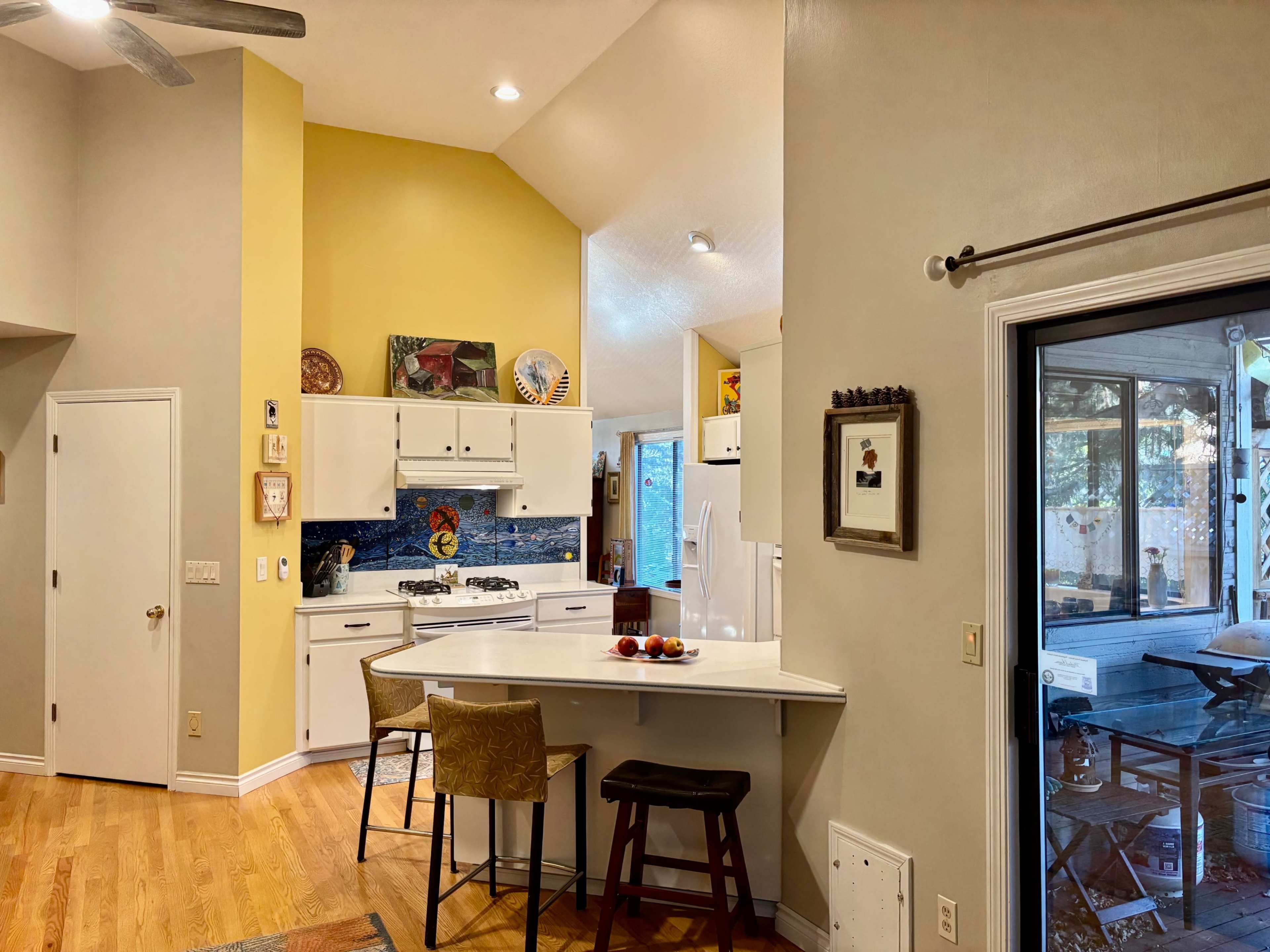The image shows a bright, modern kitchen area with white cabinets, a peninsula with two stools, and large windows that lead to a sunroom.