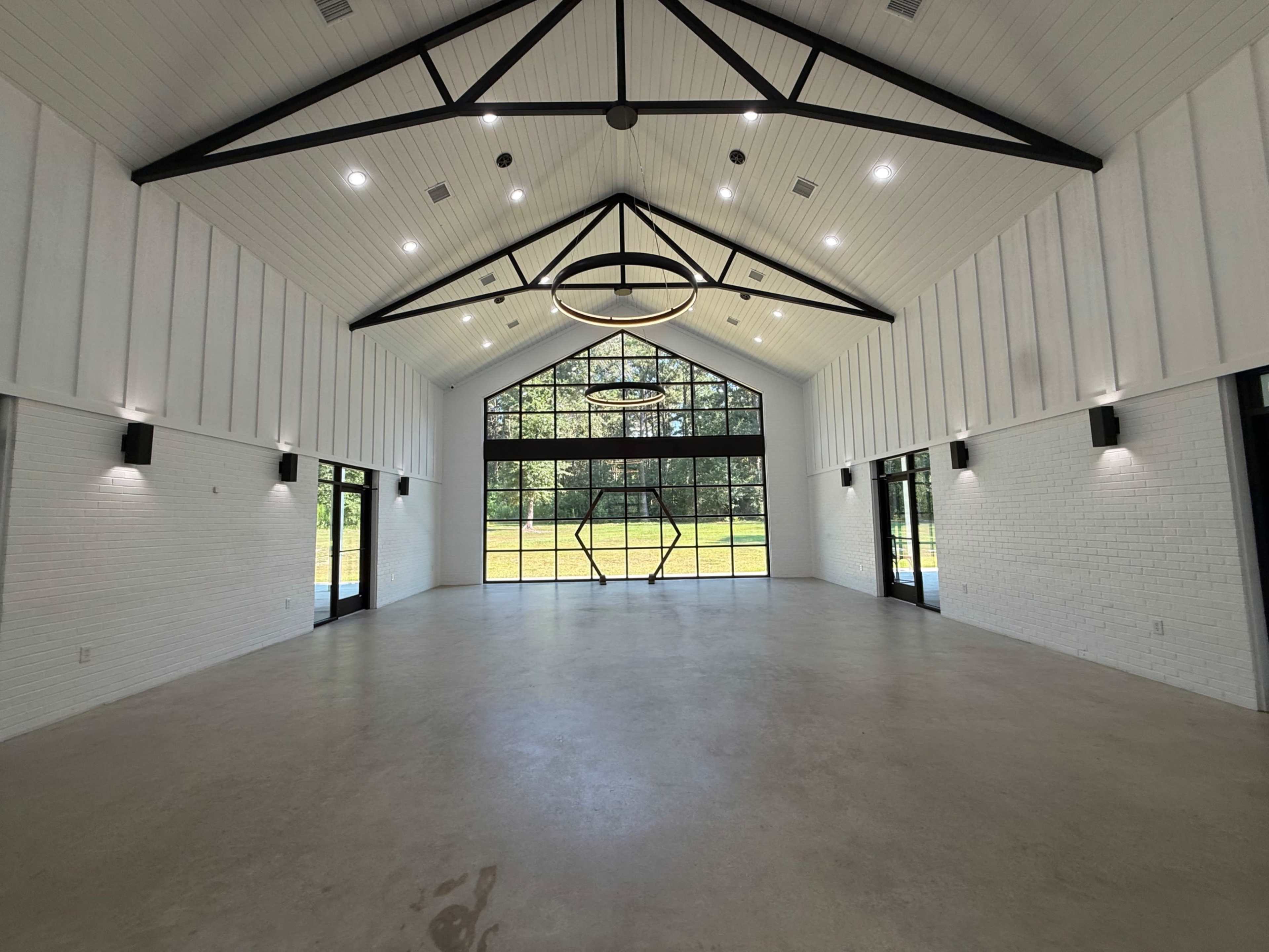 The image shows a spacious, modern interior with high ceilings, large windows, and a minimalistic design, featuring exposed beams and a neutral color palette.