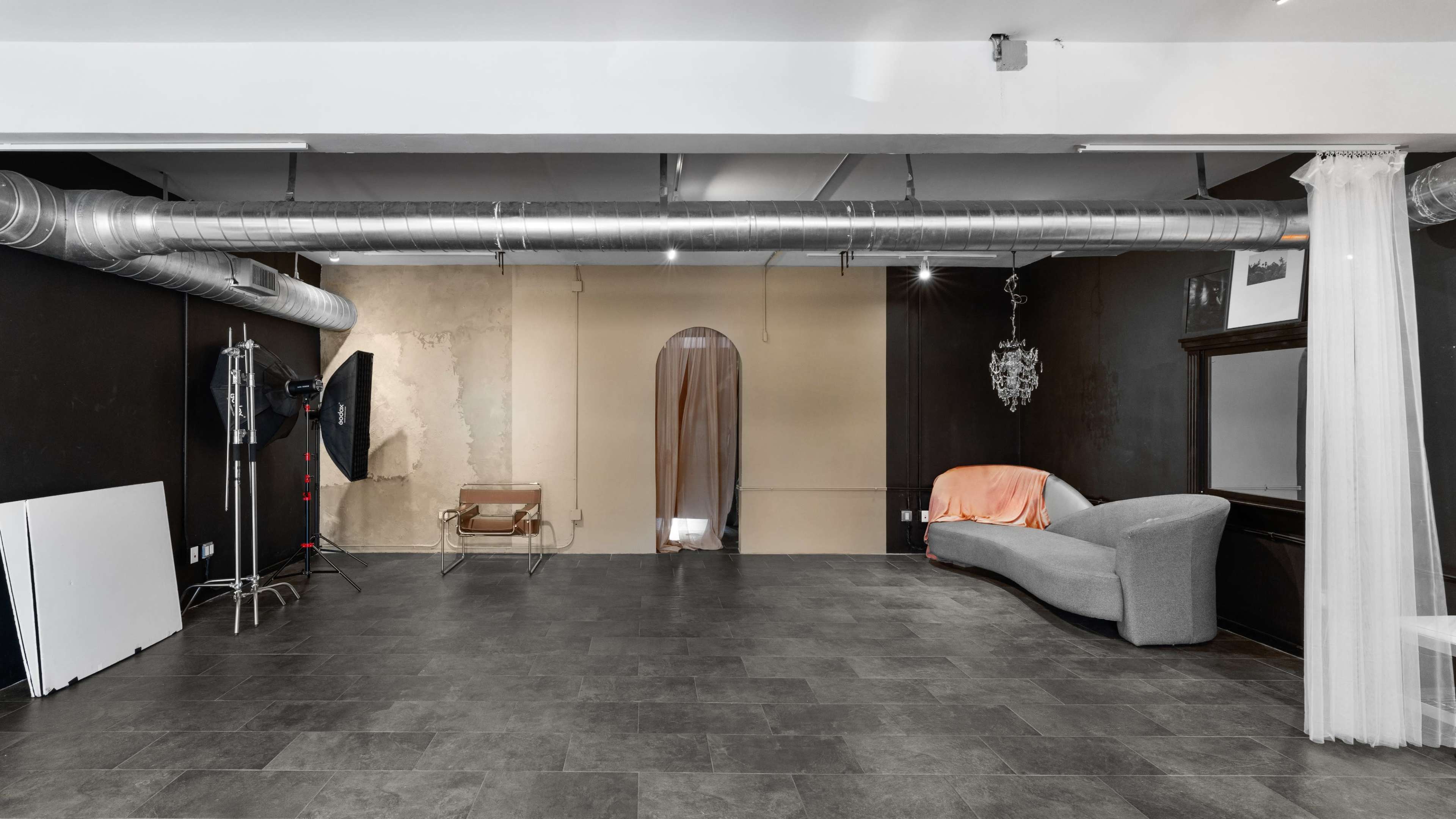 The image shows a spacious interior room with dark walls, a gray sofa, a chandelier, and large ventilation ducts overhead.