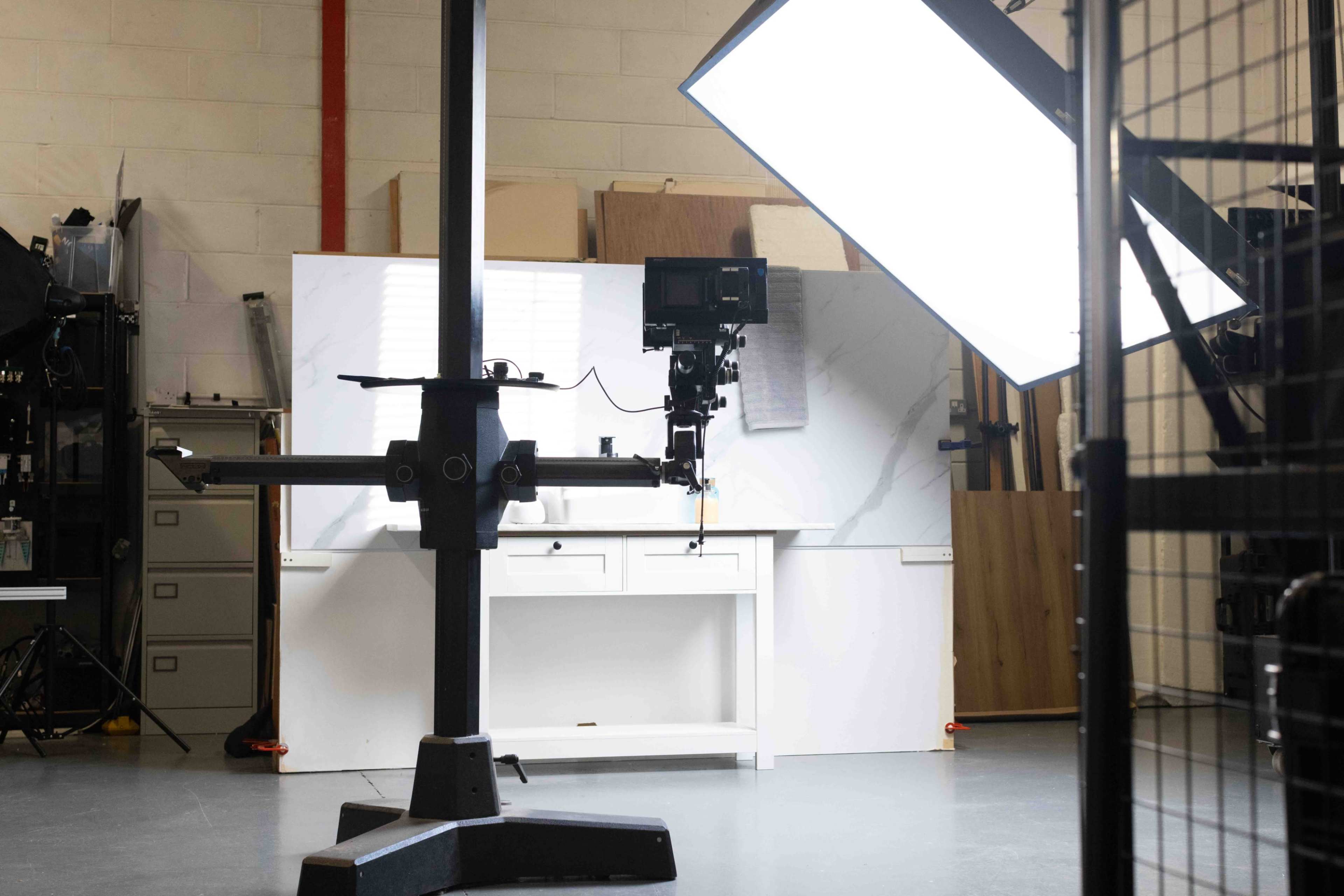The image shows a photography studio with a large lighting setup, a camera mounted on a stand, and a white backdrop with a desk.