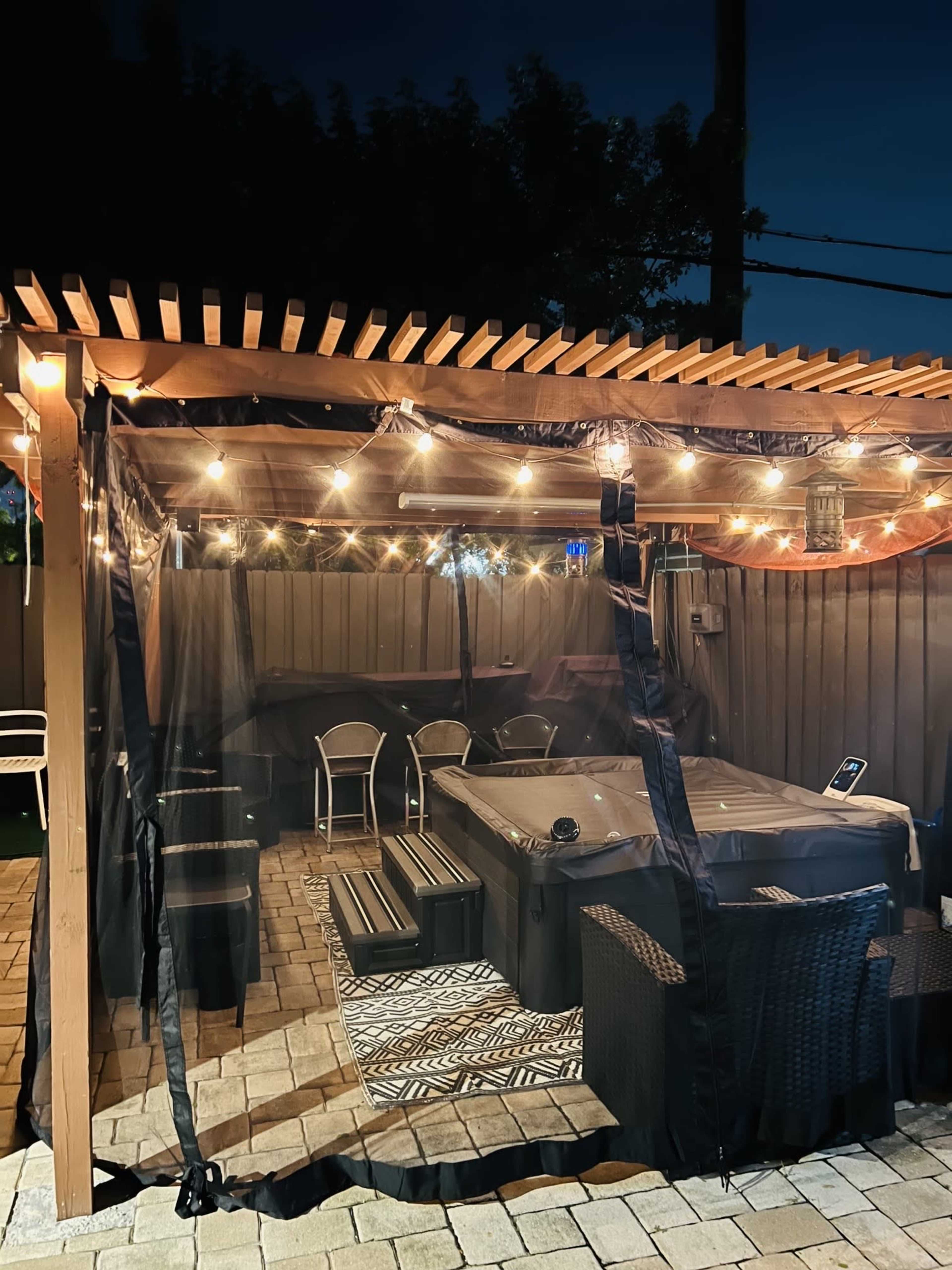 A covered outdoor space with string lights, a hot tub, and seating arrangements surrounded by a netted enclosure.