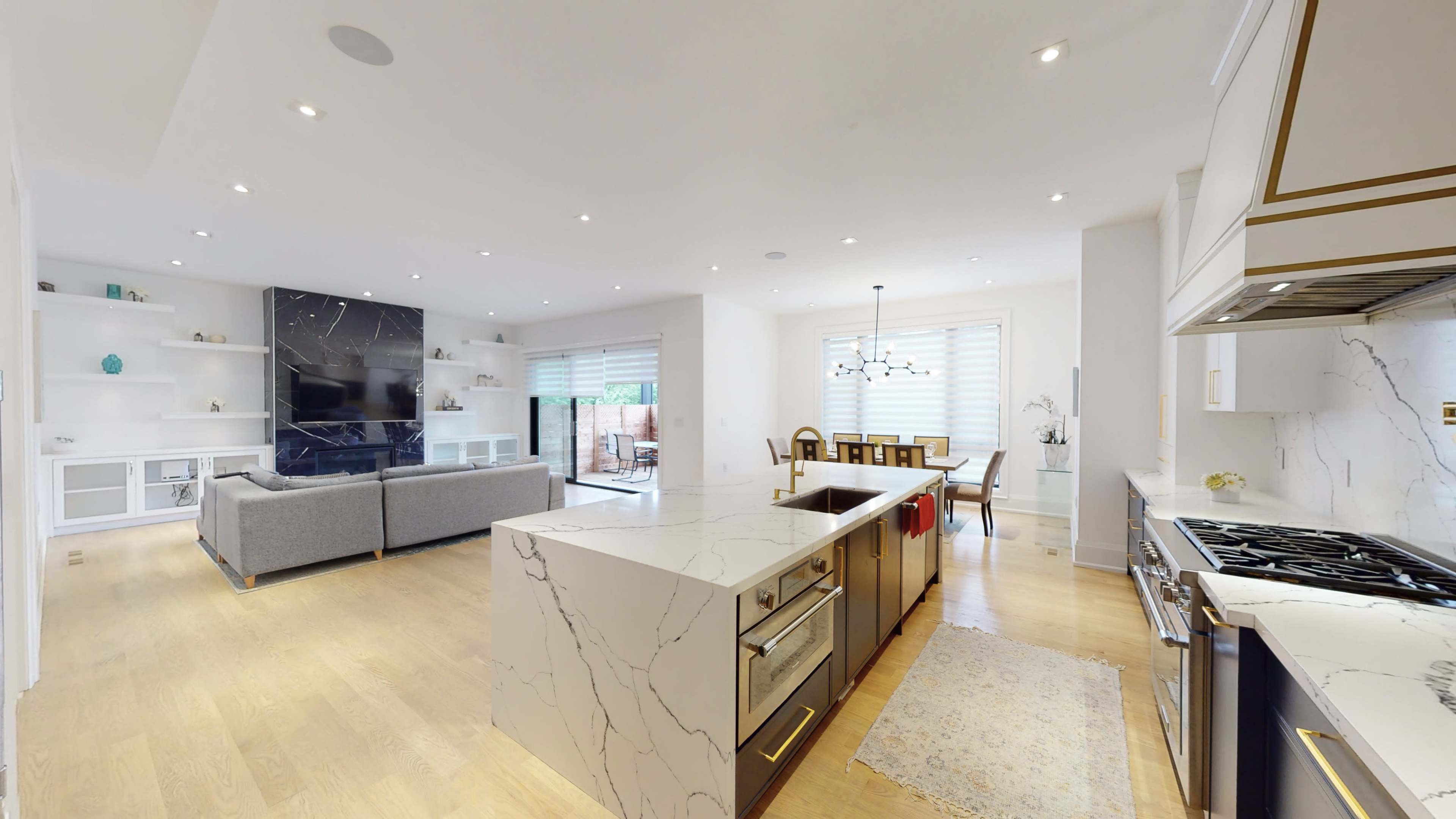 The image shows a modern kitchen and living area featuring a large island with marble countertops, a built-in fireplace, and a dining space near a sliding glass door.