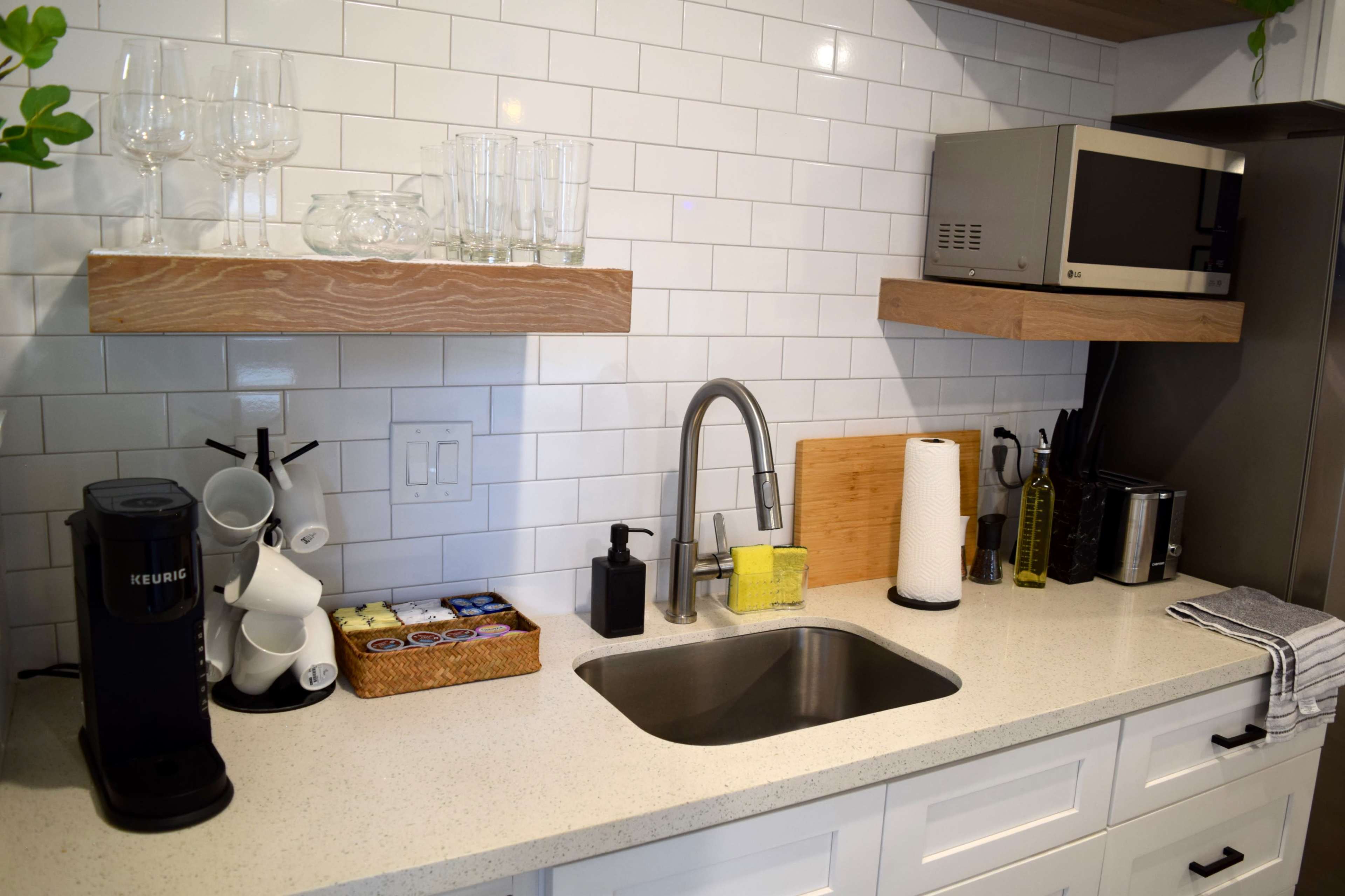 The image shows a modern kitchen countertop with a stainless steel sink, a coffee maker, a microwave, and neatly arranged utensils, all set against a white tiled backsplash.