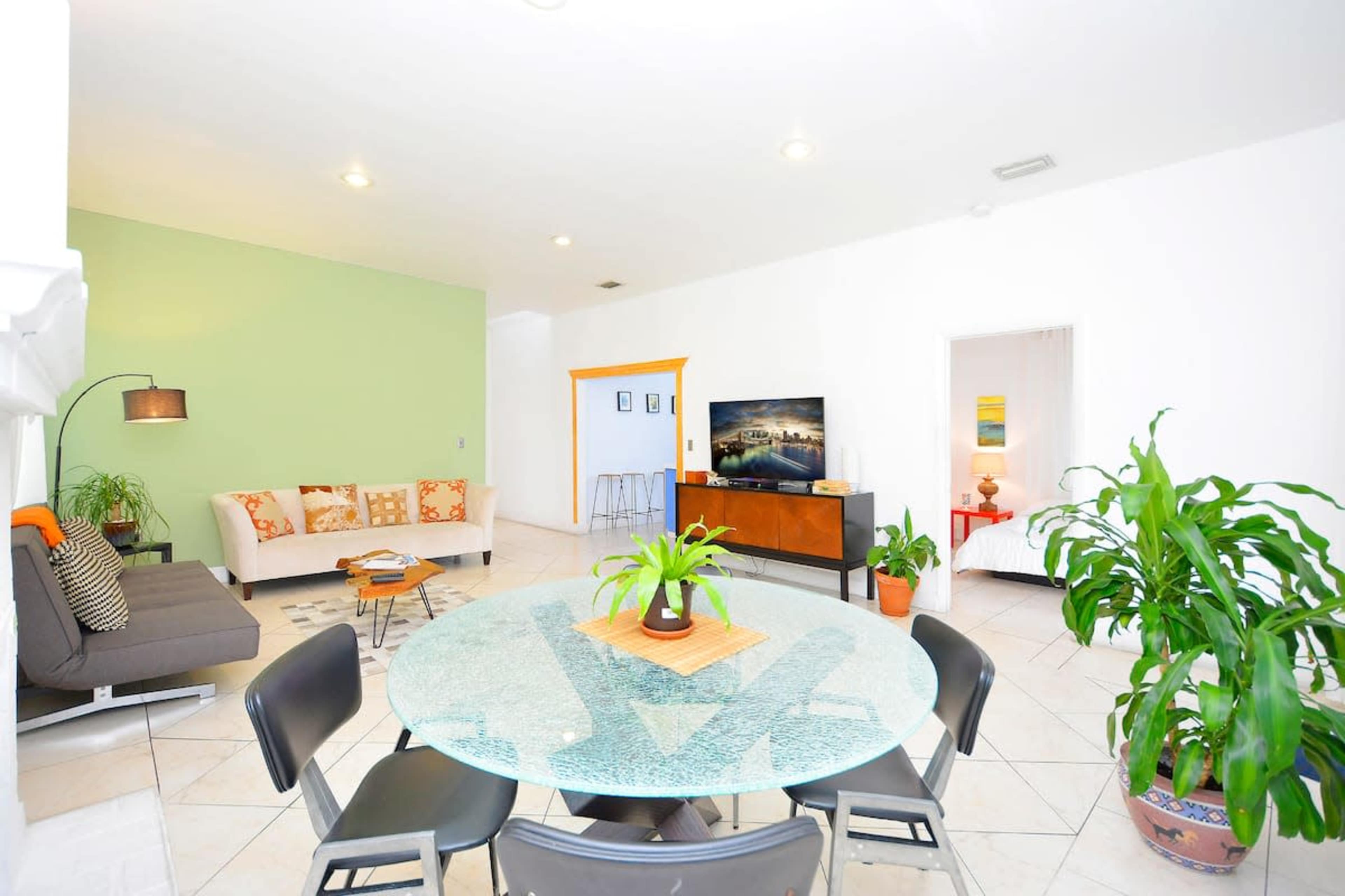 A spacious living area with a round glass dining table, black chairs, a couch with orange and white cushions, a light green accent wall, and a television on a wooden cabinet.