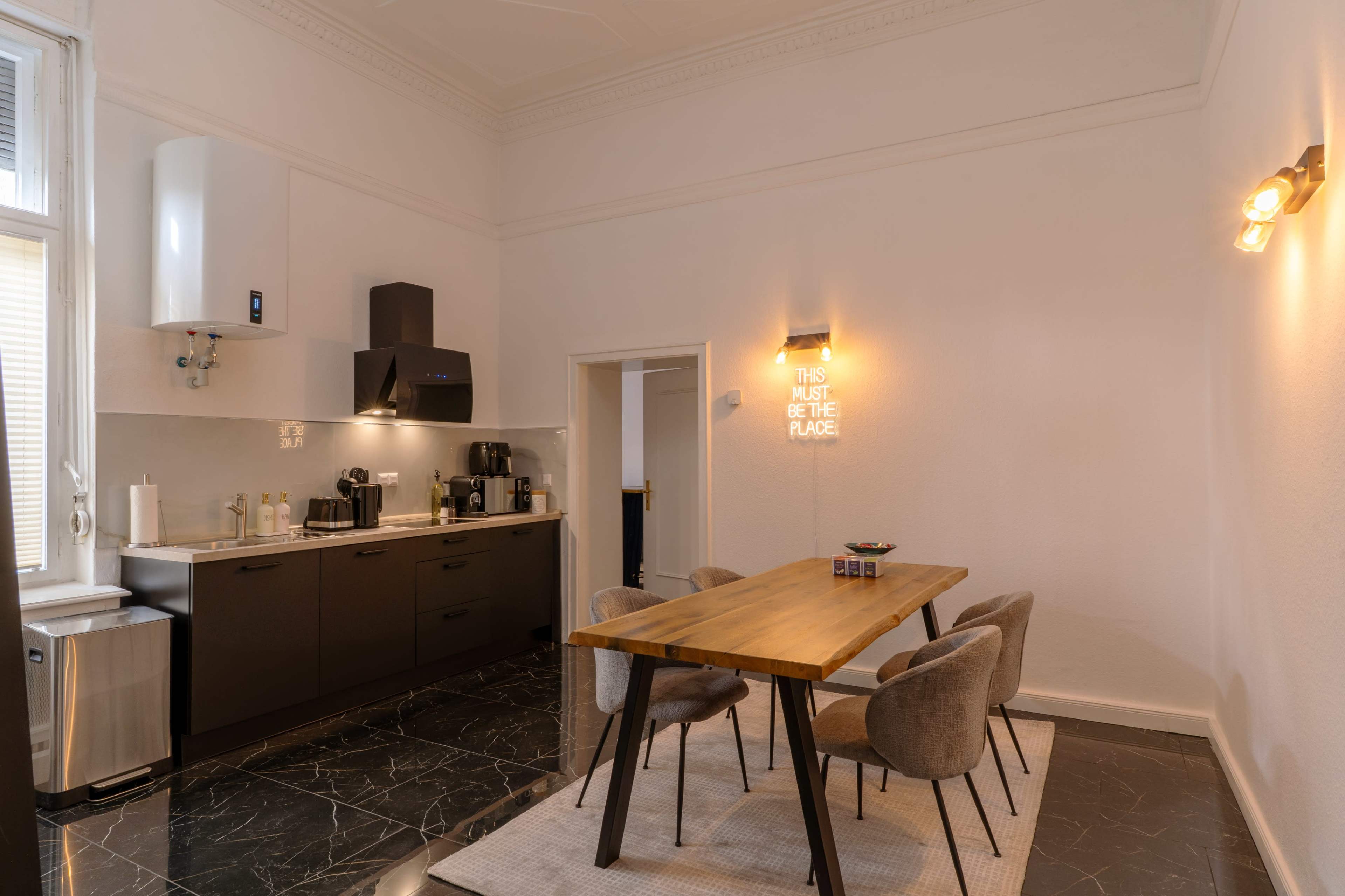 A modern kitchen features dark cabinetry, stainless steel appliances, a wooden dining table with four chairs, and wall art that reads "THIS MUST BE THE PLACE."