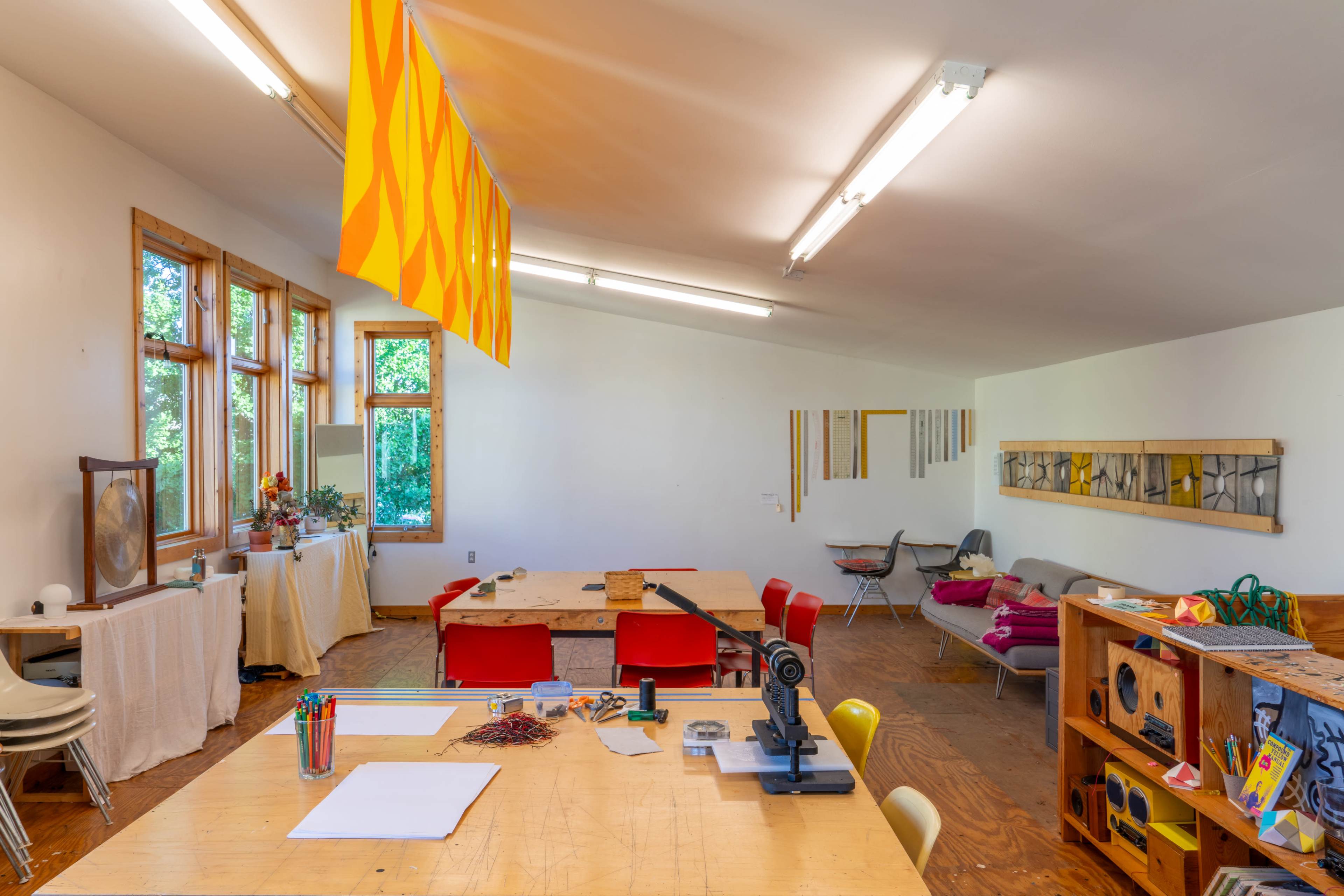 A spacious art studio features a large wooden table, colorful chairs, and windows that allow natural light to illuminate the workspace.