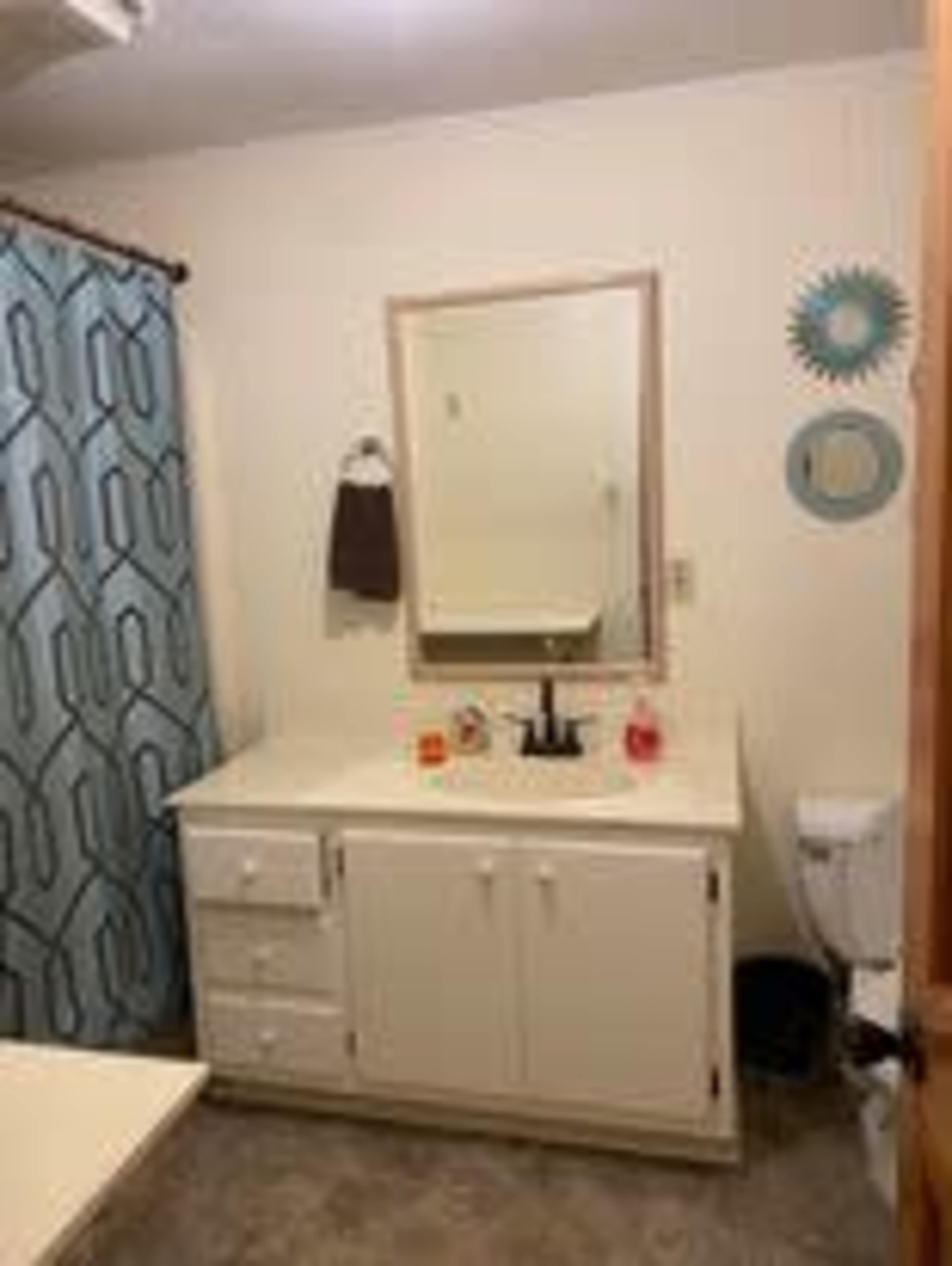 The image shows a bathroom with a white vanity, a mirror above the sink, and a shower curtain with a blue geometric pattern.