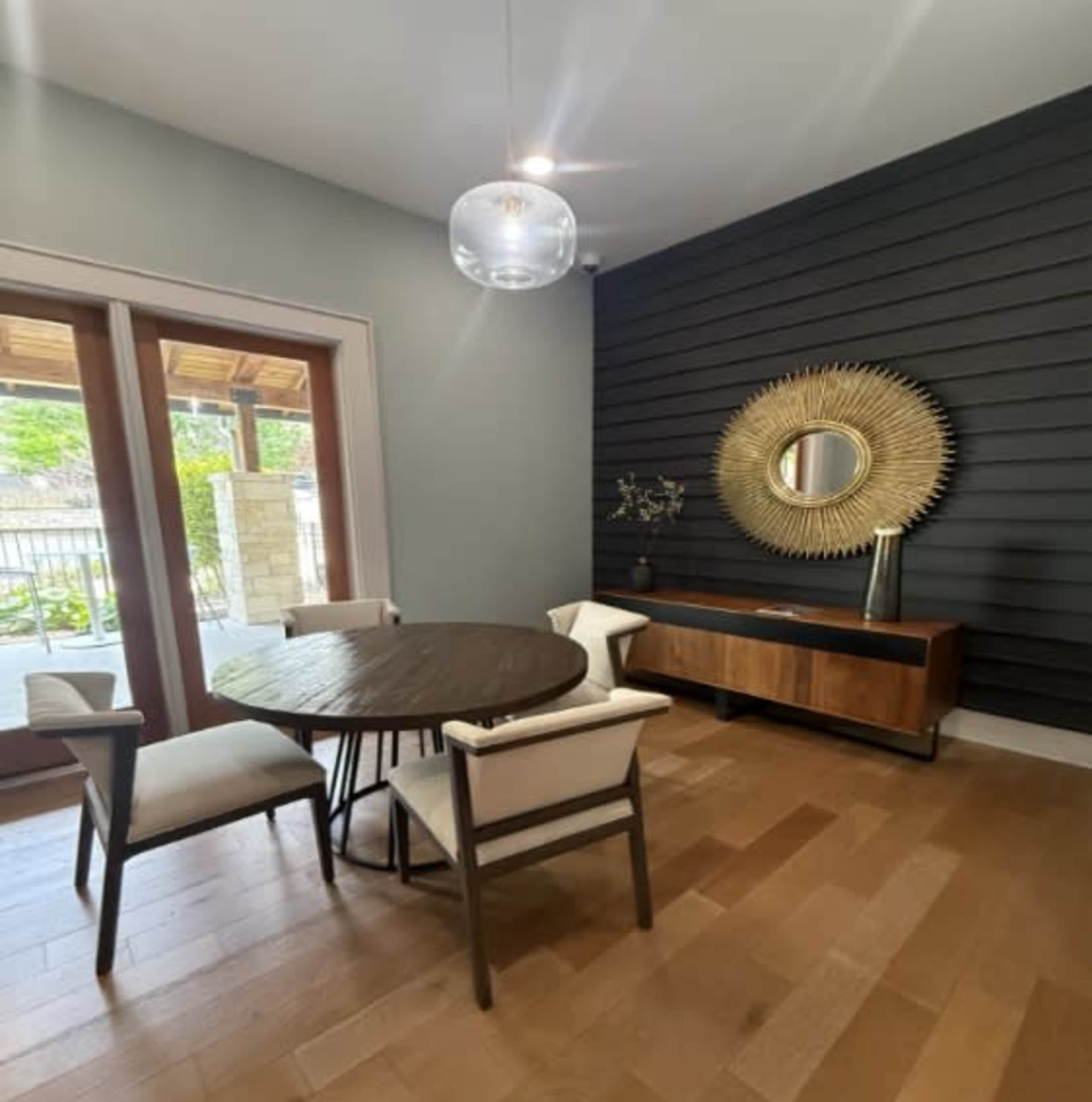 A round wooden dining table and four chairs are positioned in a modern dining area with a mirrored wall decoration and a sideboard.
