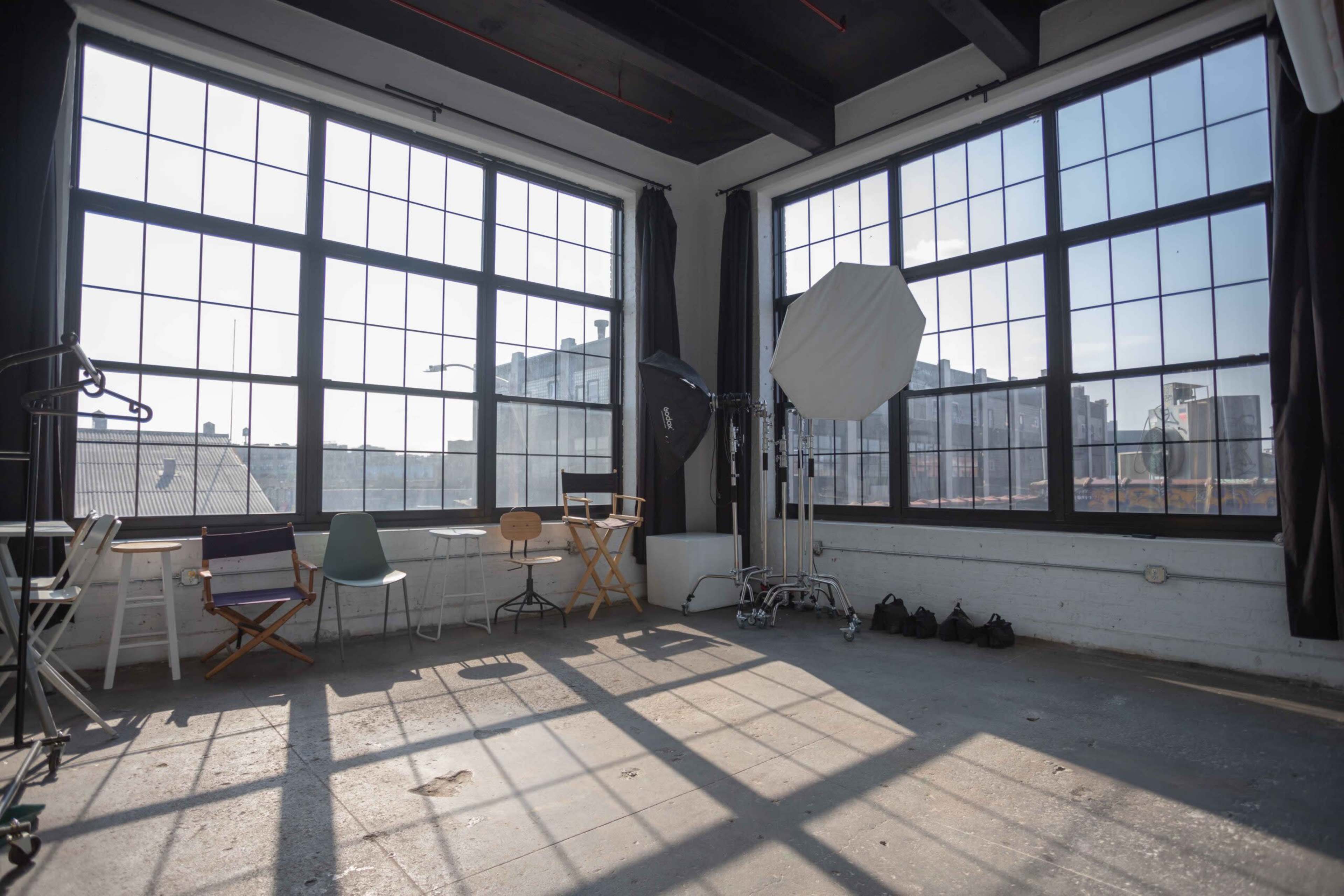 The image shows a bright, open studio space with large windows, wooden floors, and various chairs arranged around the room.