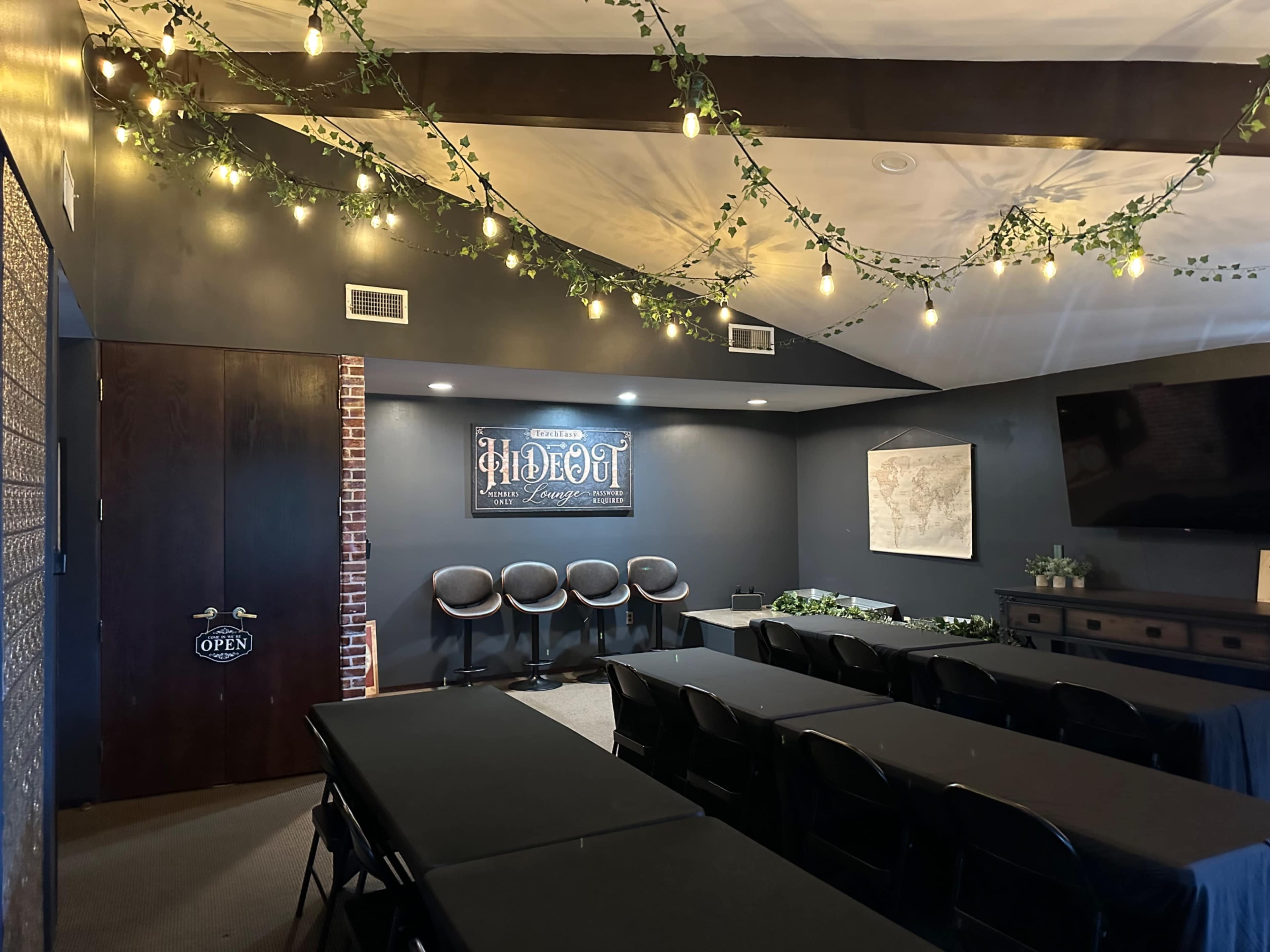 A conference room setup with black tables and chairs, decorated with string lights and greenery, featuring a wall sign that reads "Hideout" and a TV on one side.