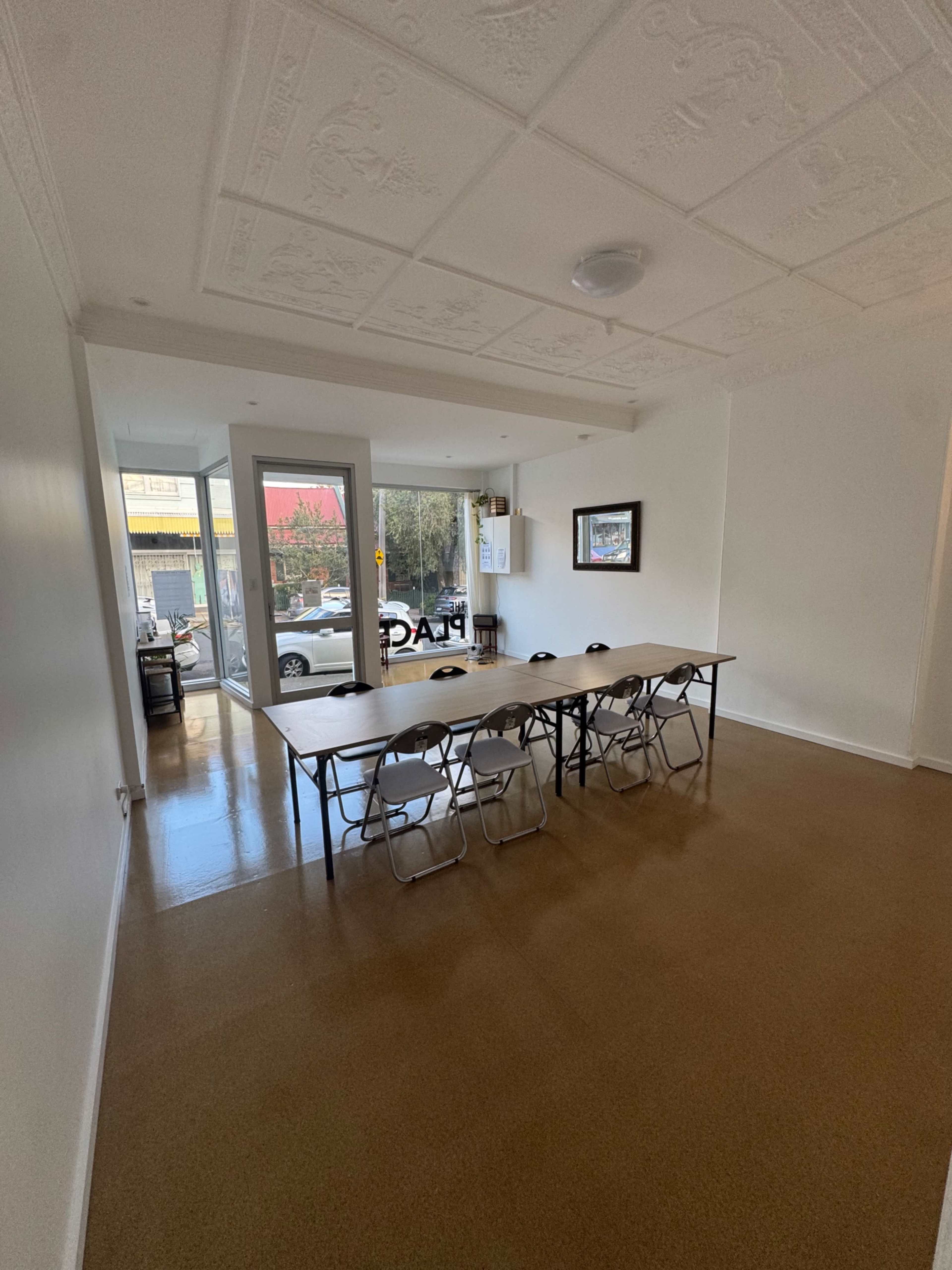 The image shows a bright room featuring a long table with several chairs arranged around it, leading to a window that overlooks a street.