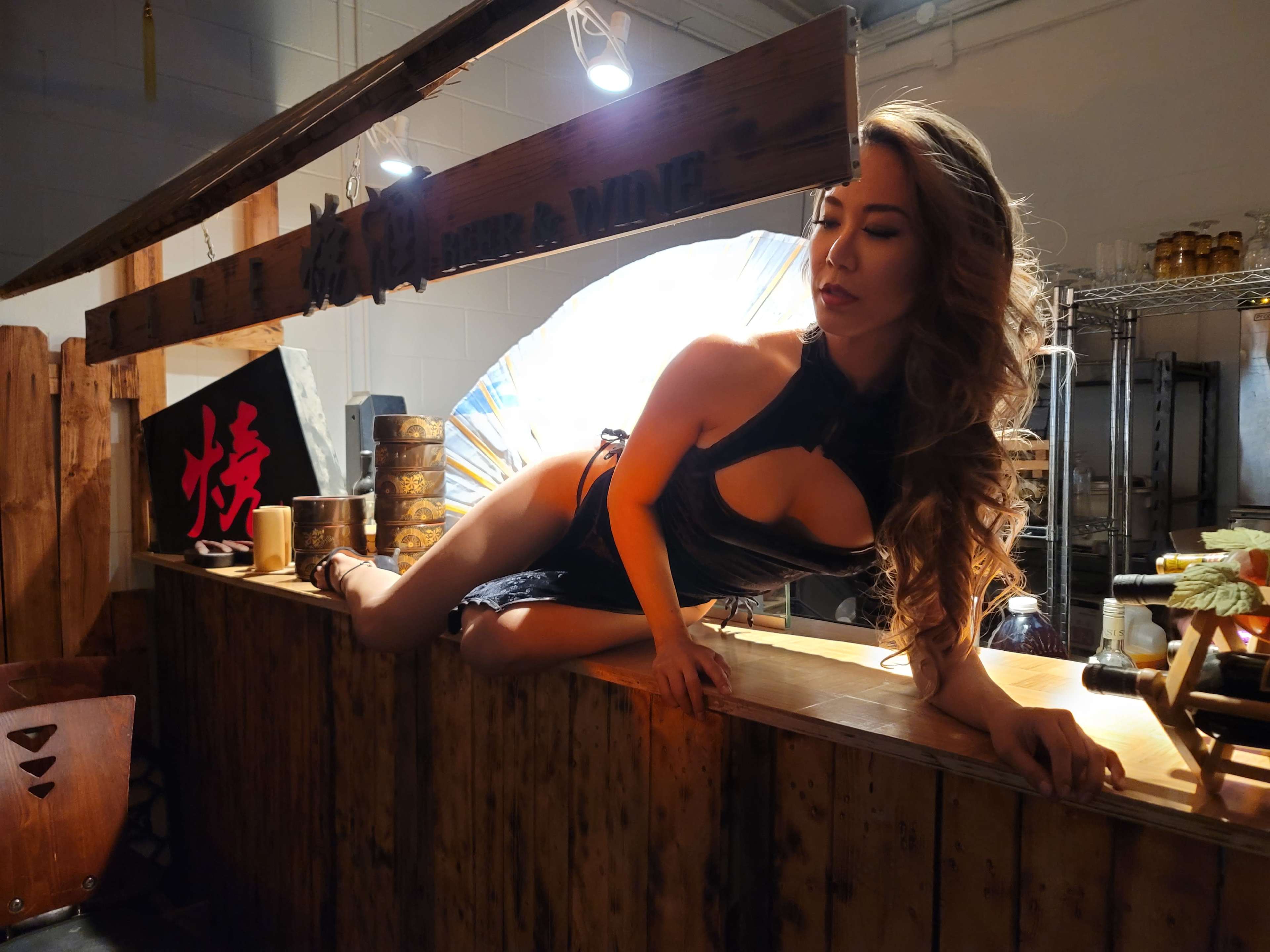 A woman poses on a wooden bar counter in an indoor setting decorated with Asian-inspired elements.